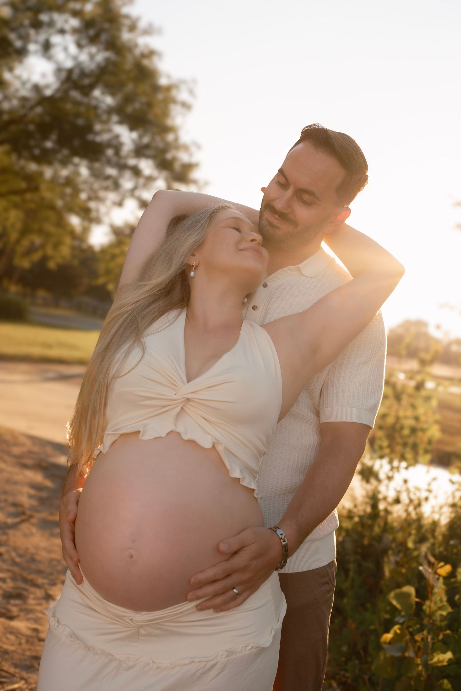 A couple poses outdoors at golden hour, the pregnant woman cradling her baby bump while her partner embraces her from behind.