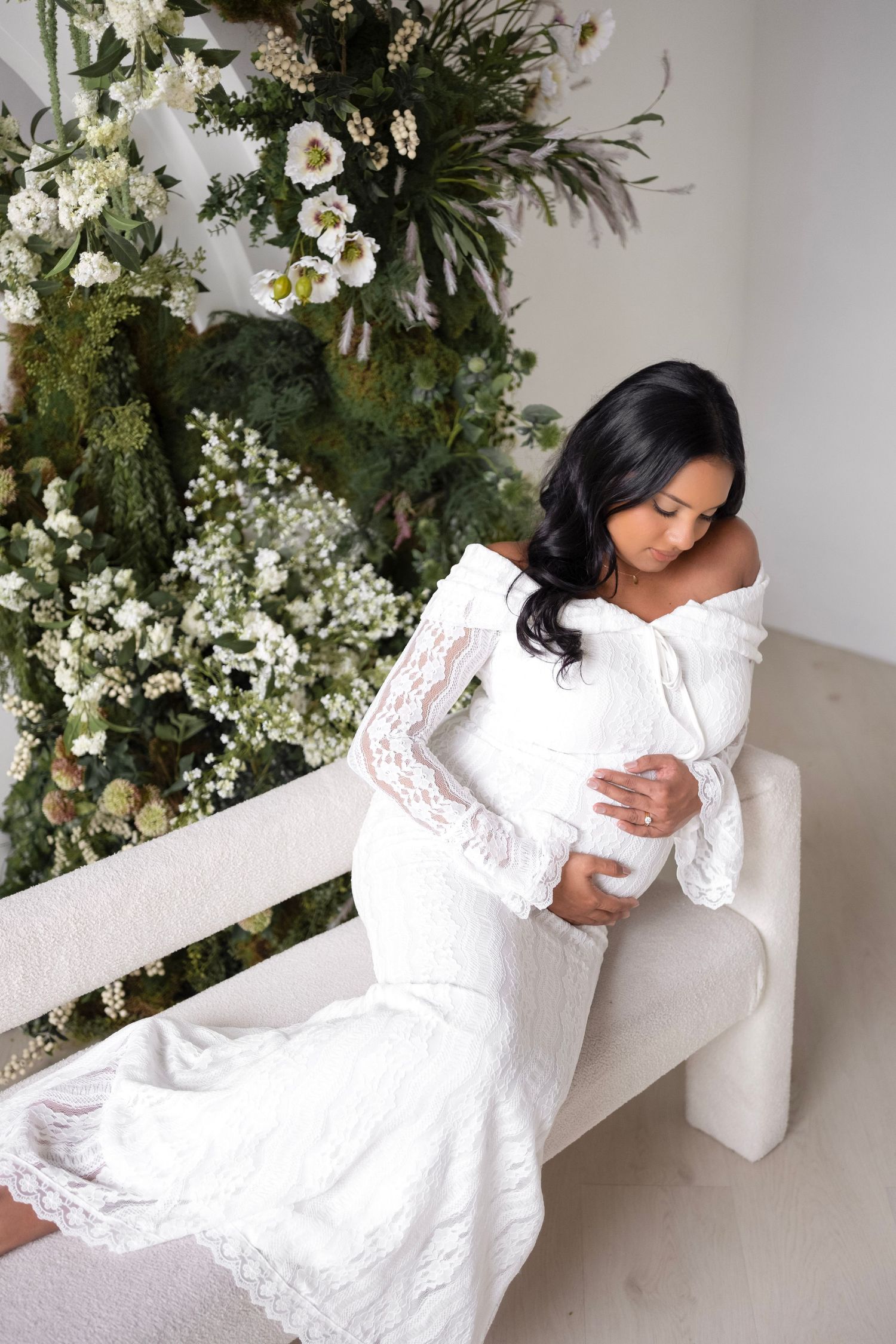 Pregnant woman in white lace off-shoulder gown sits on bench, cradling baby bump, surrounded by lush floral backdrop.