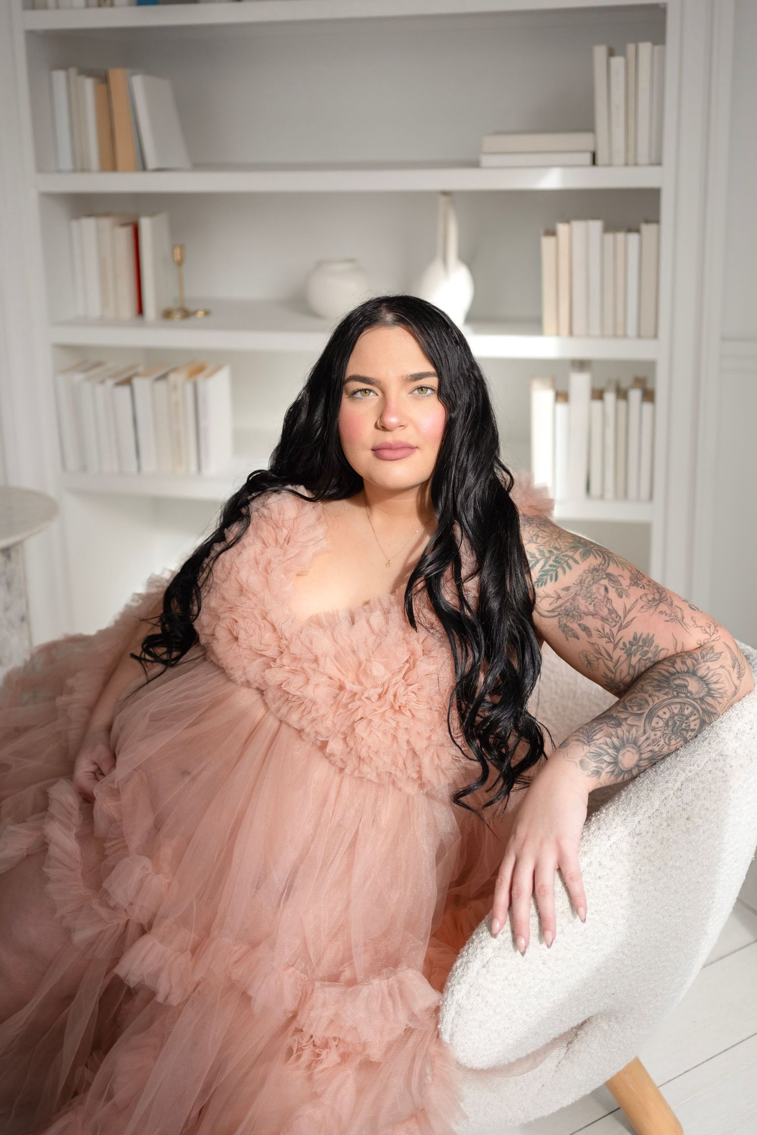 A woman in a pink tulle dress with tattoos sits elegantly in a white chair before a bookshelf.
