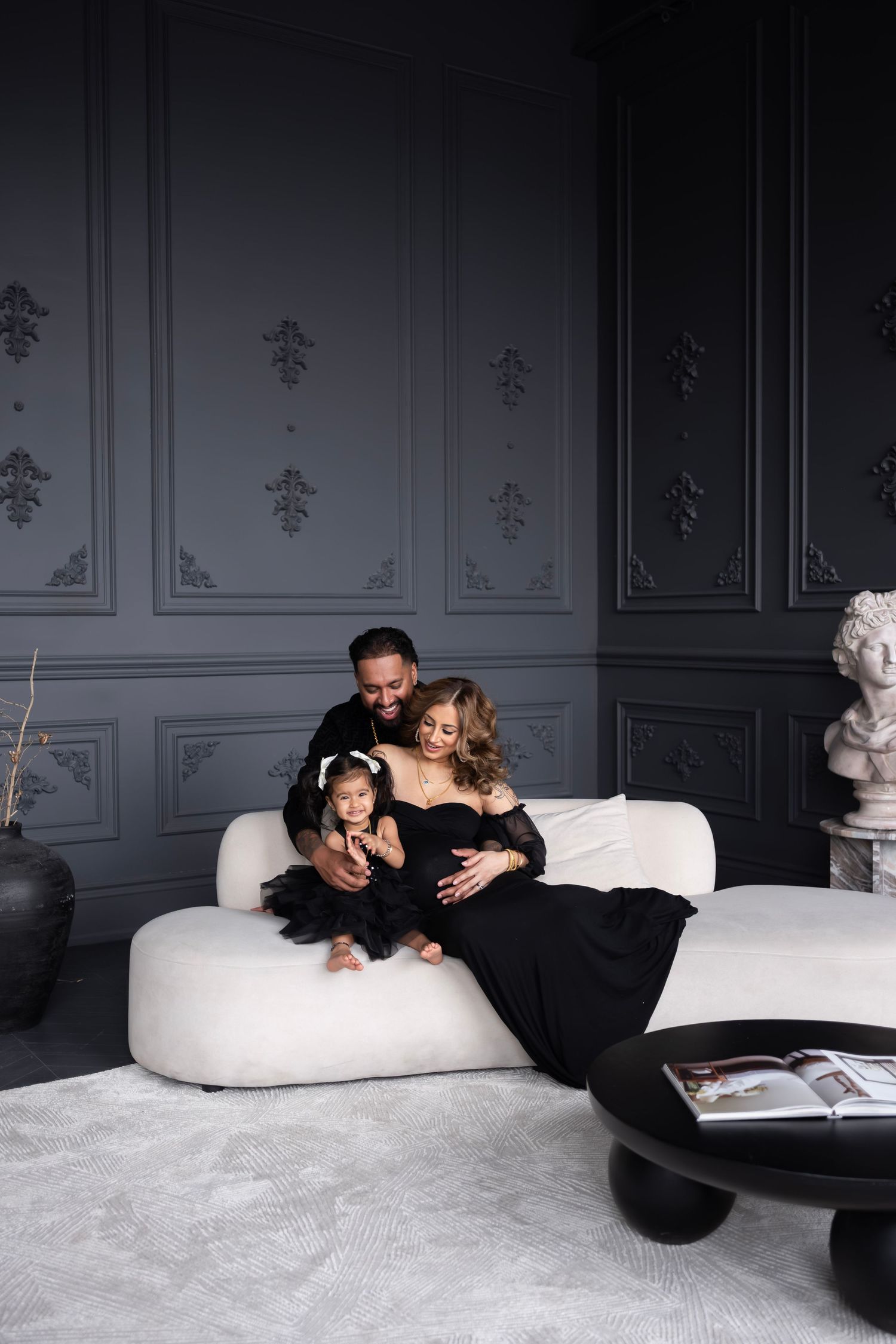 Family in matching black outfits sitting on white sofa, pregnant mother with husband and toddler daughter.