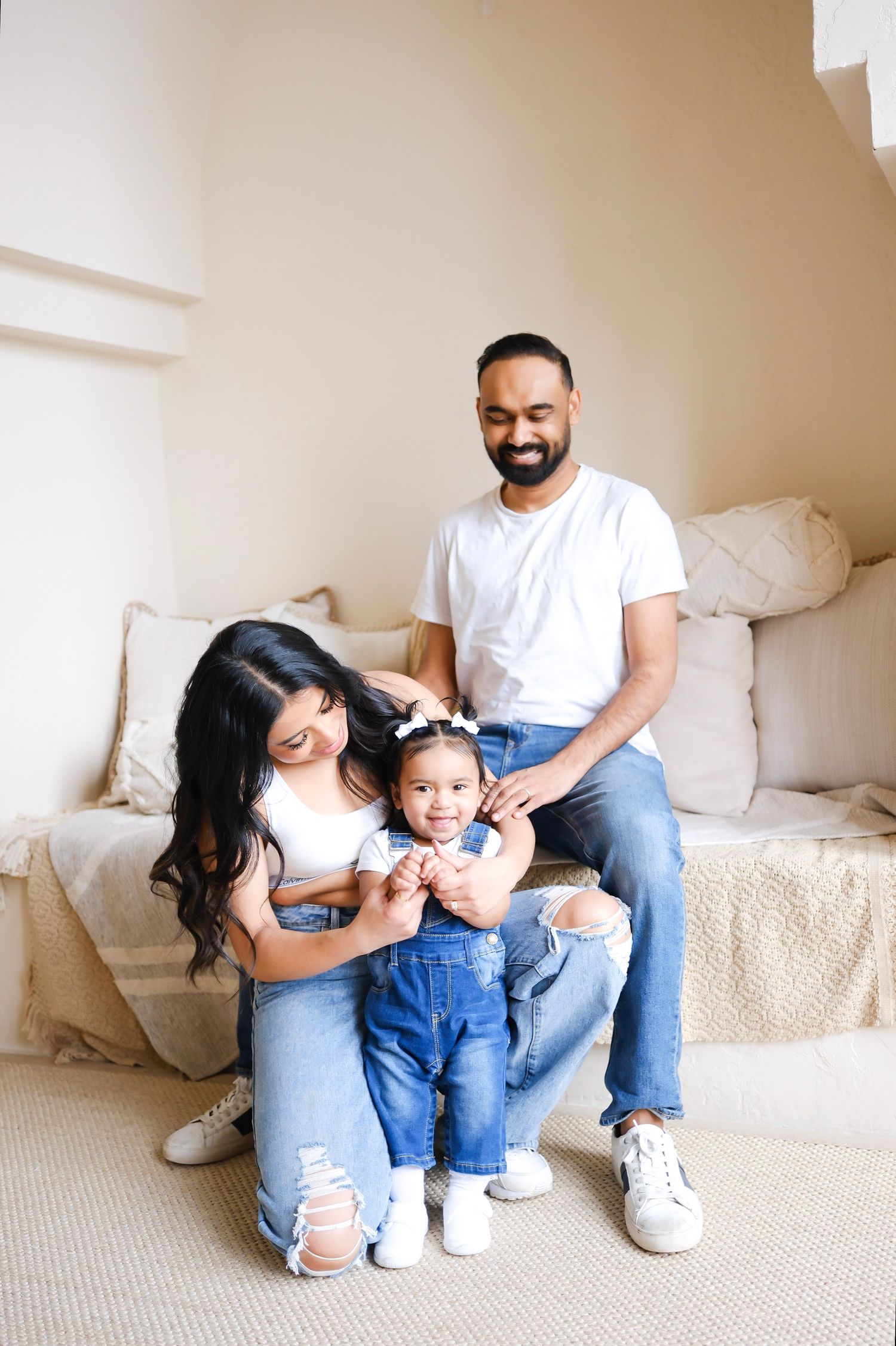 A young family sits cozily together on beige carpet against neutral walls in matching casual denim and white outfits.