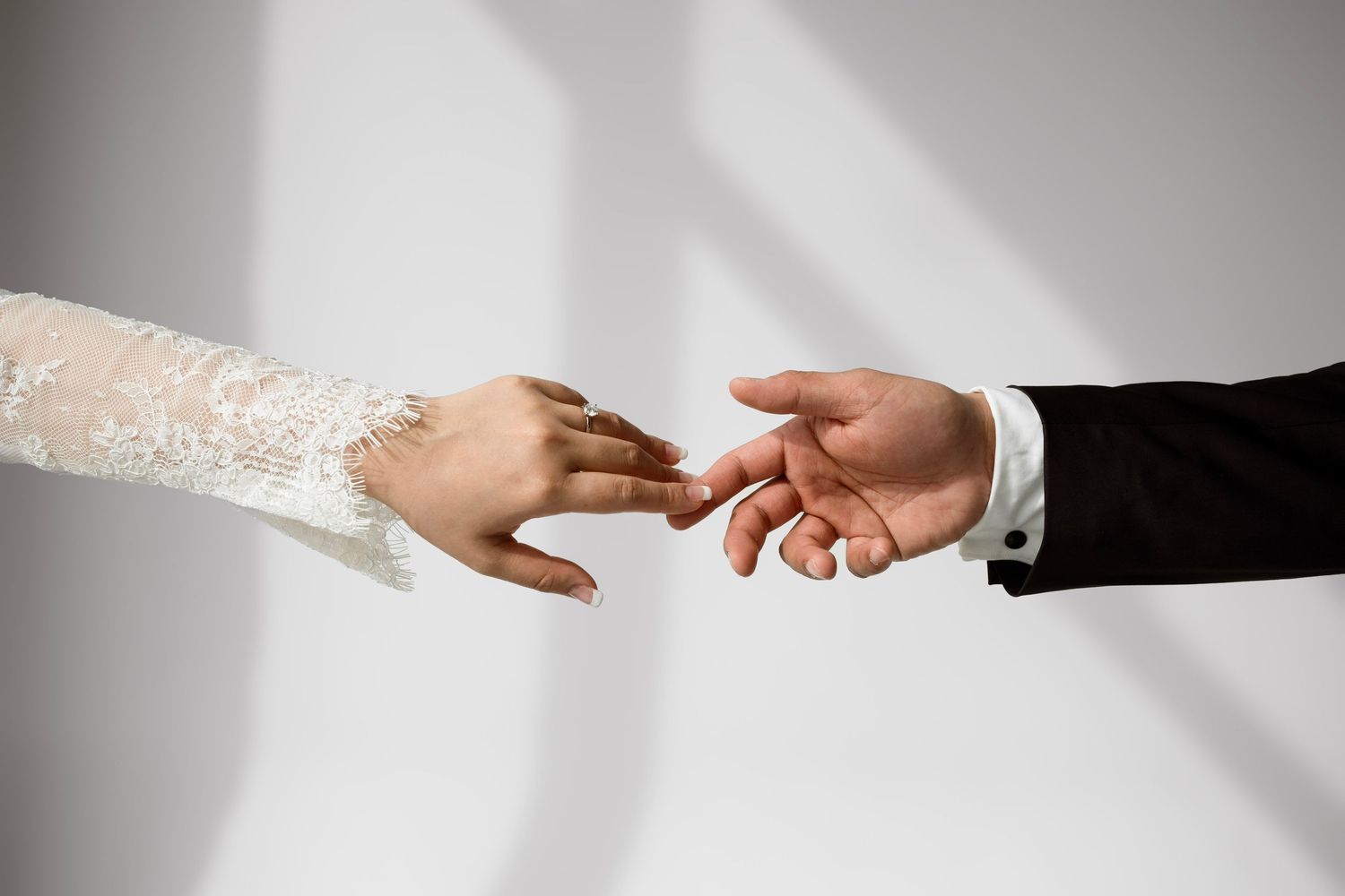 Bride and groom reaching hands toward each other, showcasing wedding attire with lace sleeve and black suit.