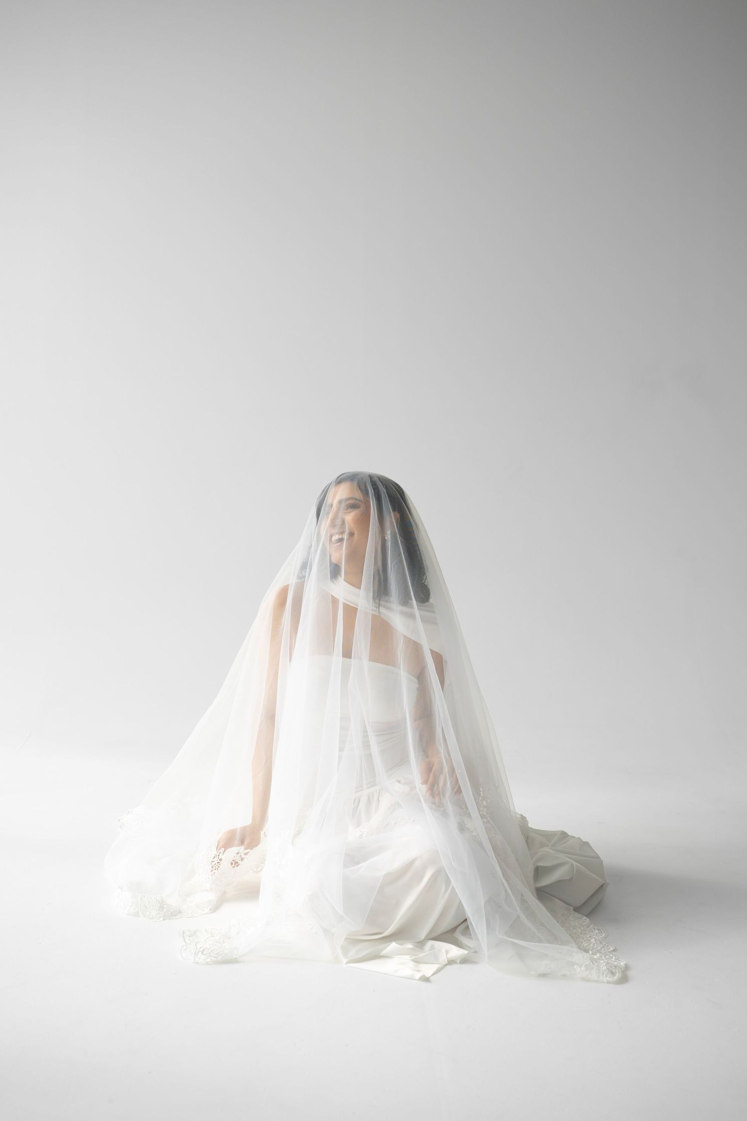 Smiling bride seated on floor with flowing lace veil draped over her head in a white studio.