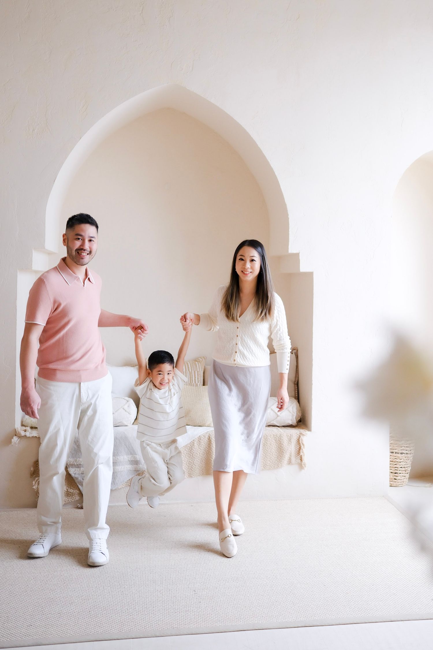 A happy family plays and jumps together in a bright white room with arched architectural details.