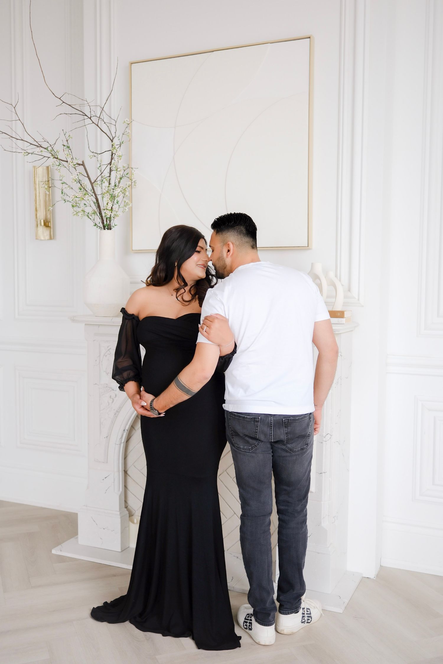 A couple in formal attire share an intimate moment in front of a minimalist white wall with decorative branches.