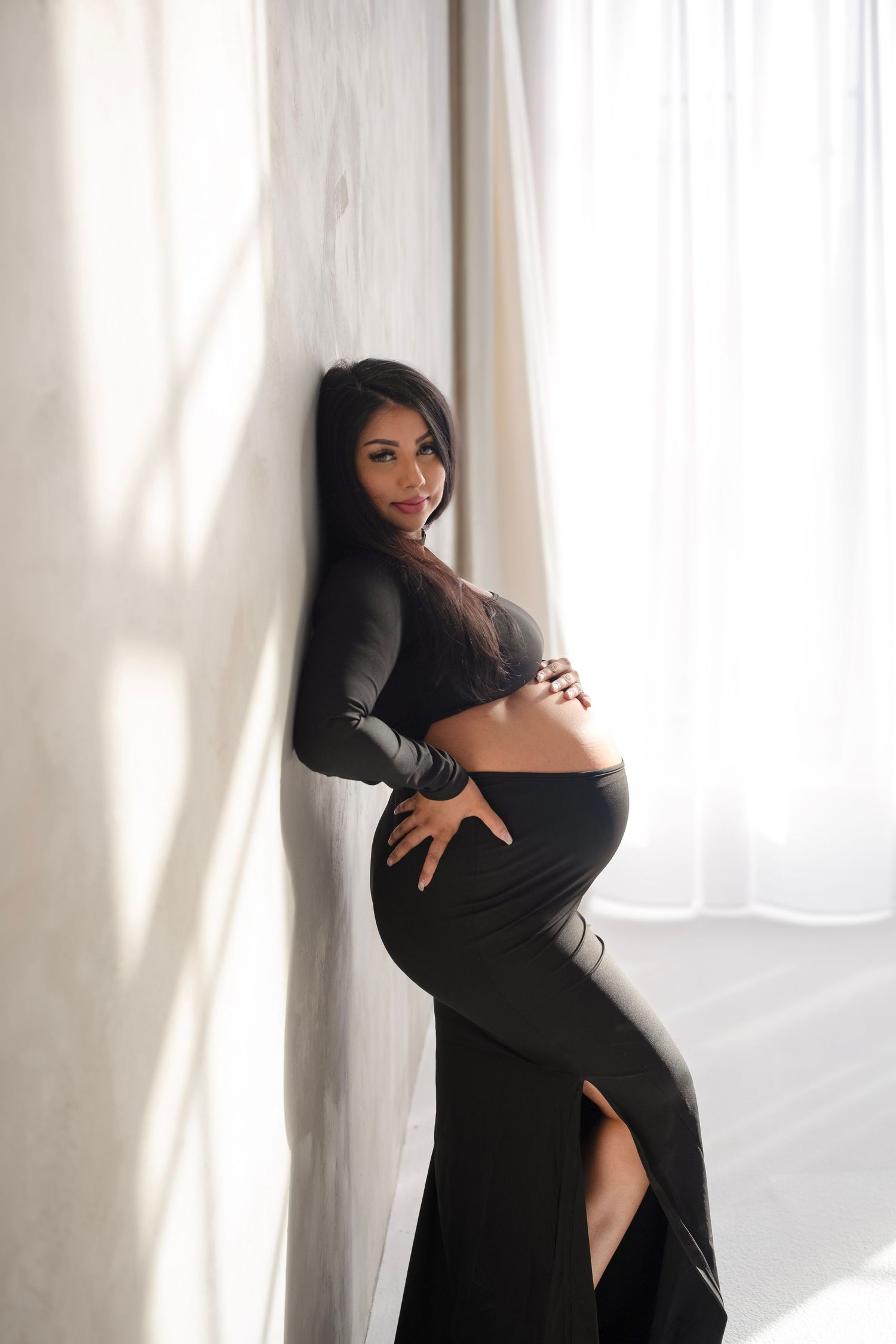 A silhouetted figure in black poses gracefully against white curtains in a minimalist boudoir photoshoot.