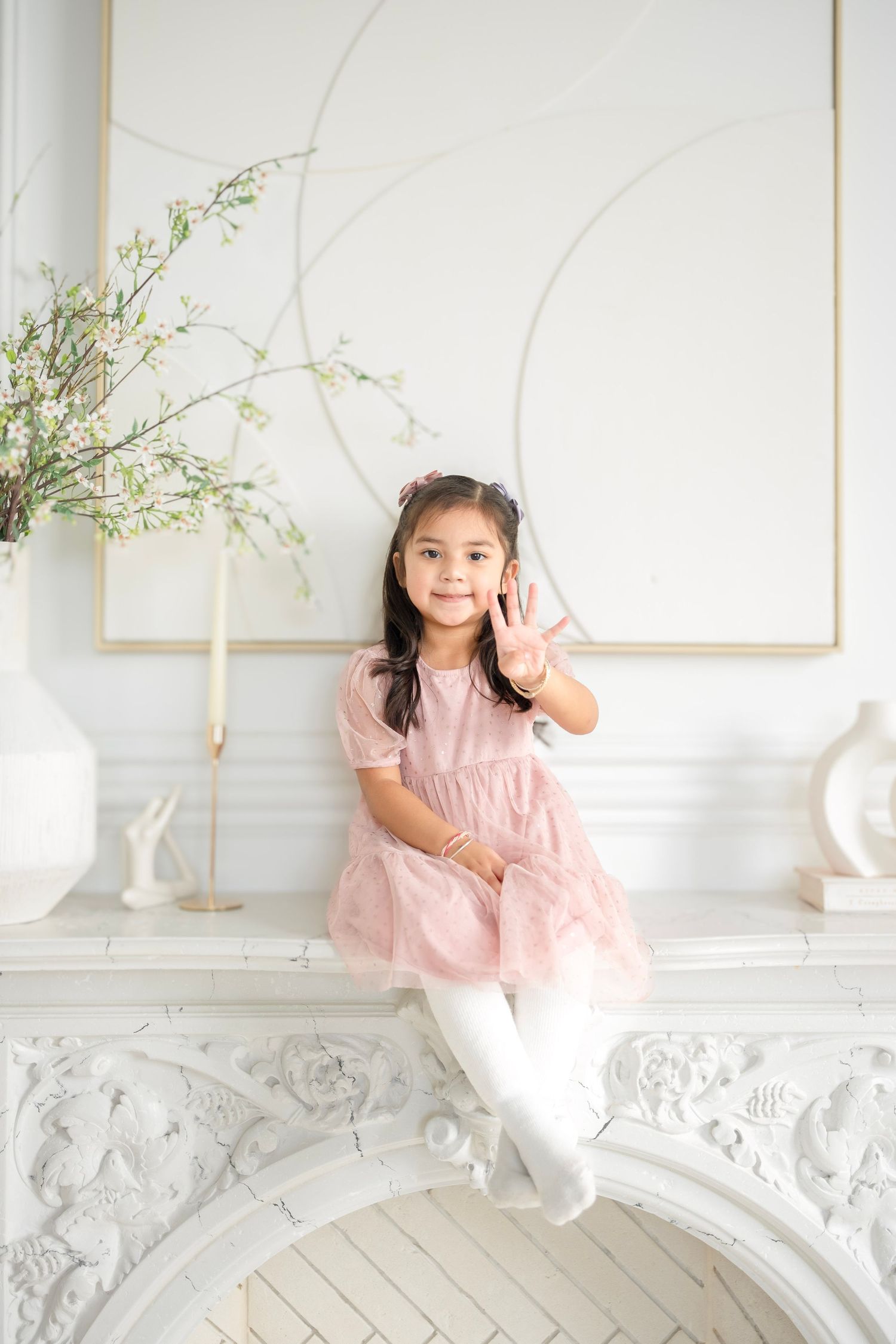 Young girl in pink tulle dress sitting on white ornate fireplace mantel, holding up three fingers and smiling.