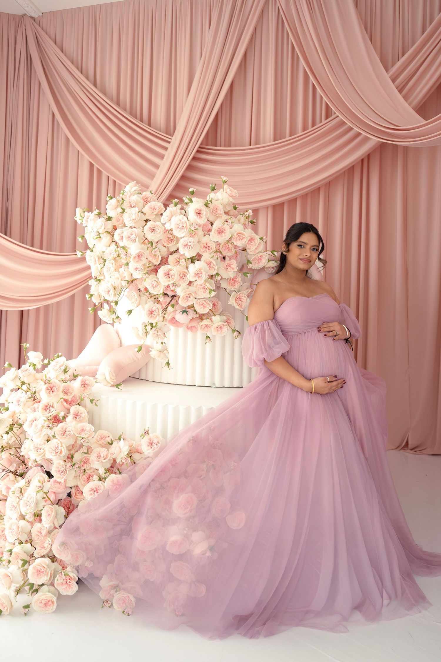 A dreamy maternity photoshoot featuring a flowing lilac gown against a pink draped backdrop with white floral arrangements.
