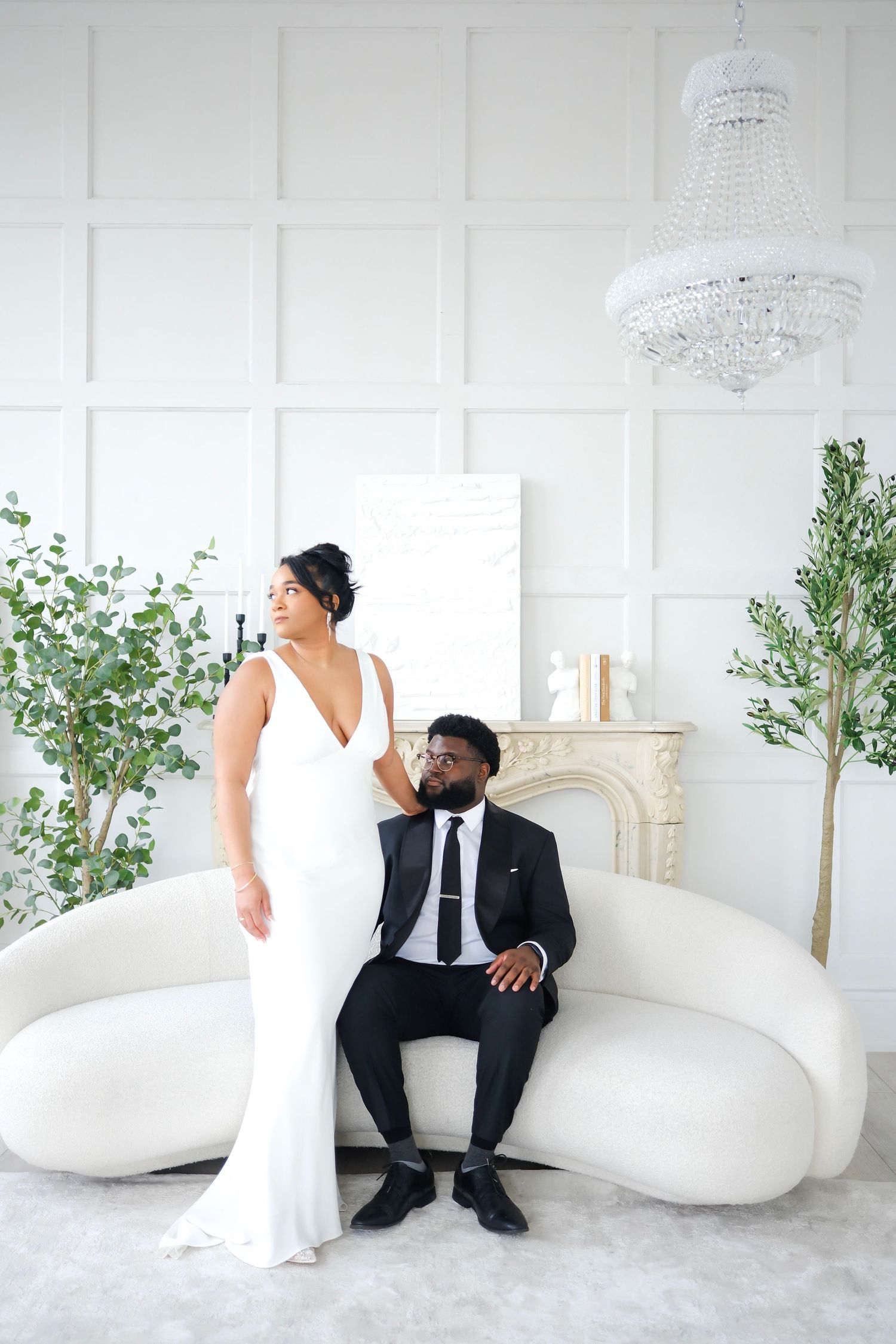 Elegant newlyweds pose in all-white room with crystal chandelier and greenery, bride in sleek gown and groom in black suit.