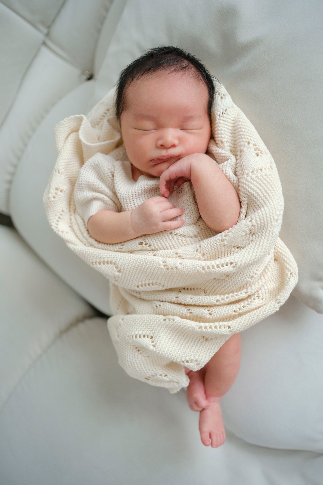 Peaceful newborn baby sleeping while wrapped in a cream knitted blanket on white bedding.