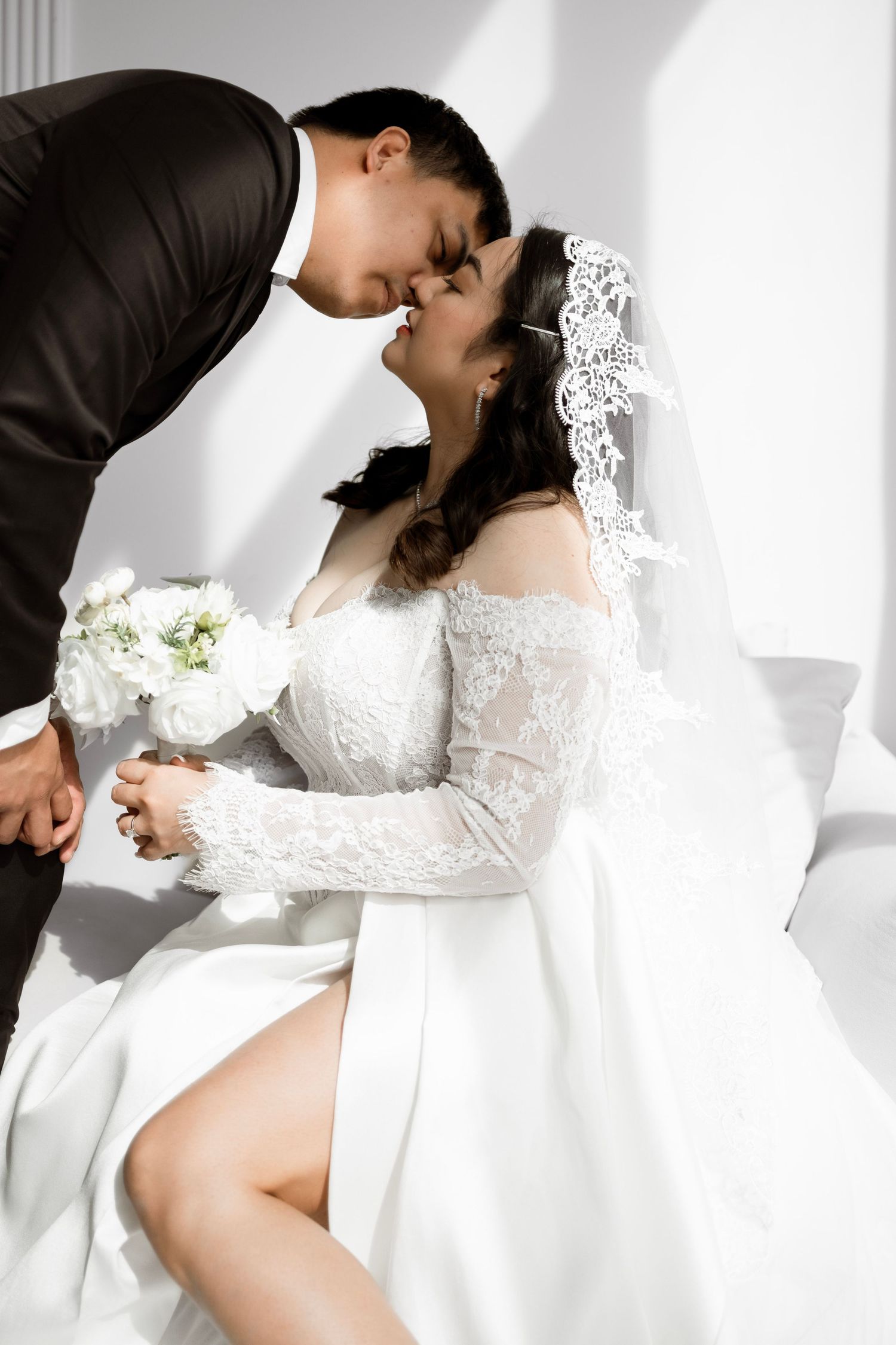 Bride in white lace off-shoulder gown with veil holds bouquet as groom in black suit leans in for a kiss.