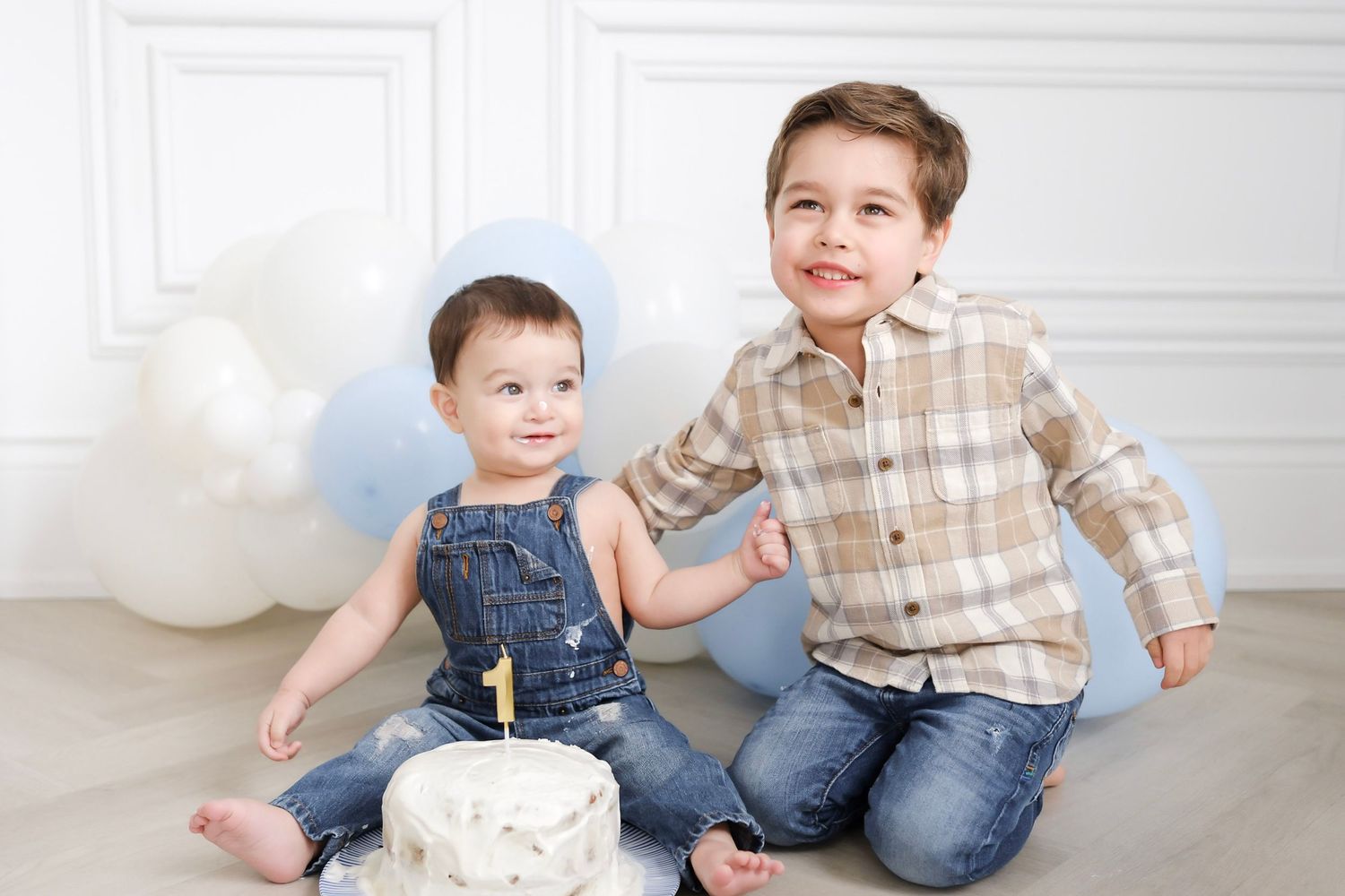 Two young boys sit together with a smash cake celebrating a first birthday, surrounded by blue and white balloons.