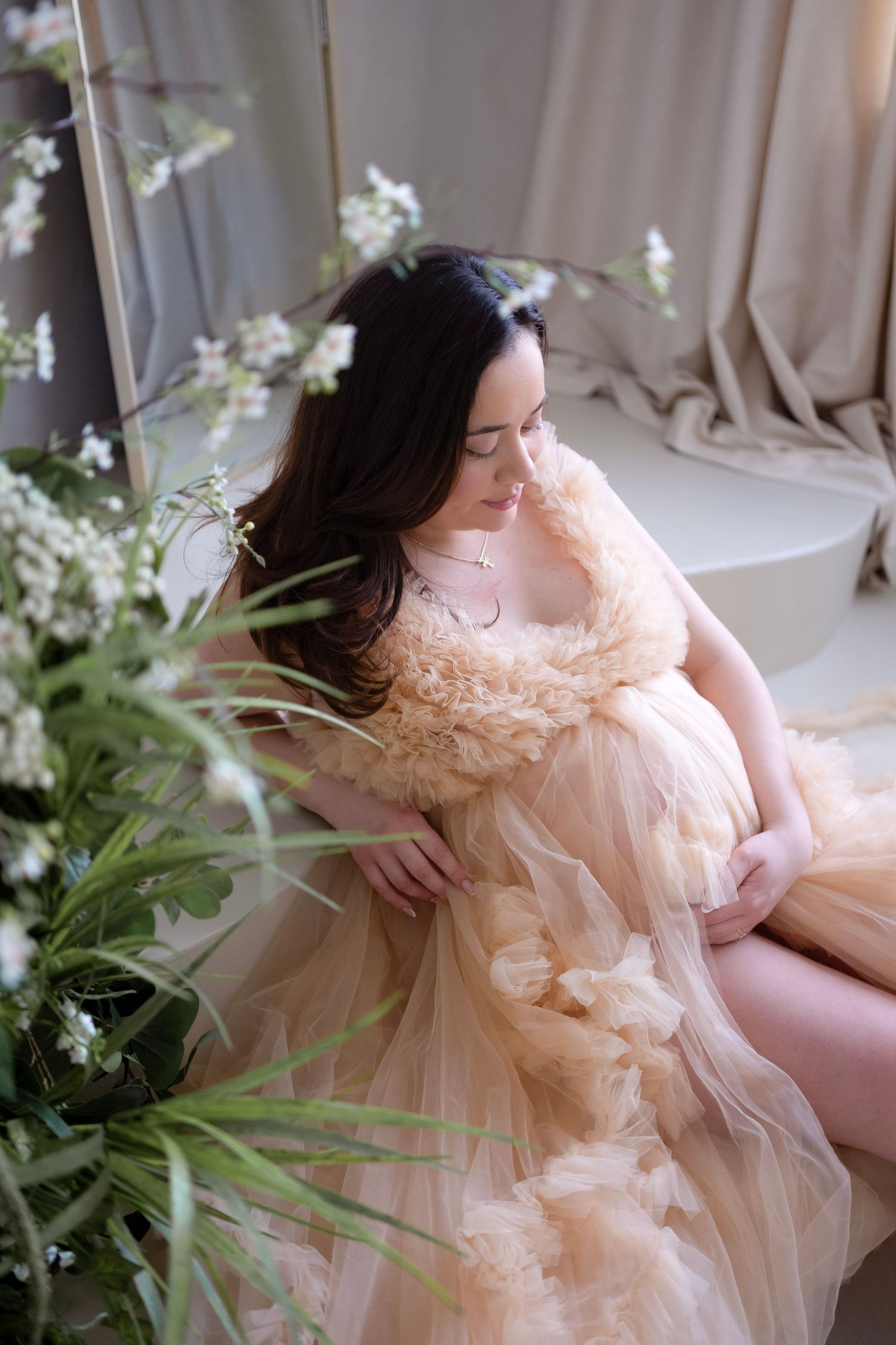 Pregnant woman in elegant peach tulle gown sitting gracefully among white flowers in a softly lit studio setting.