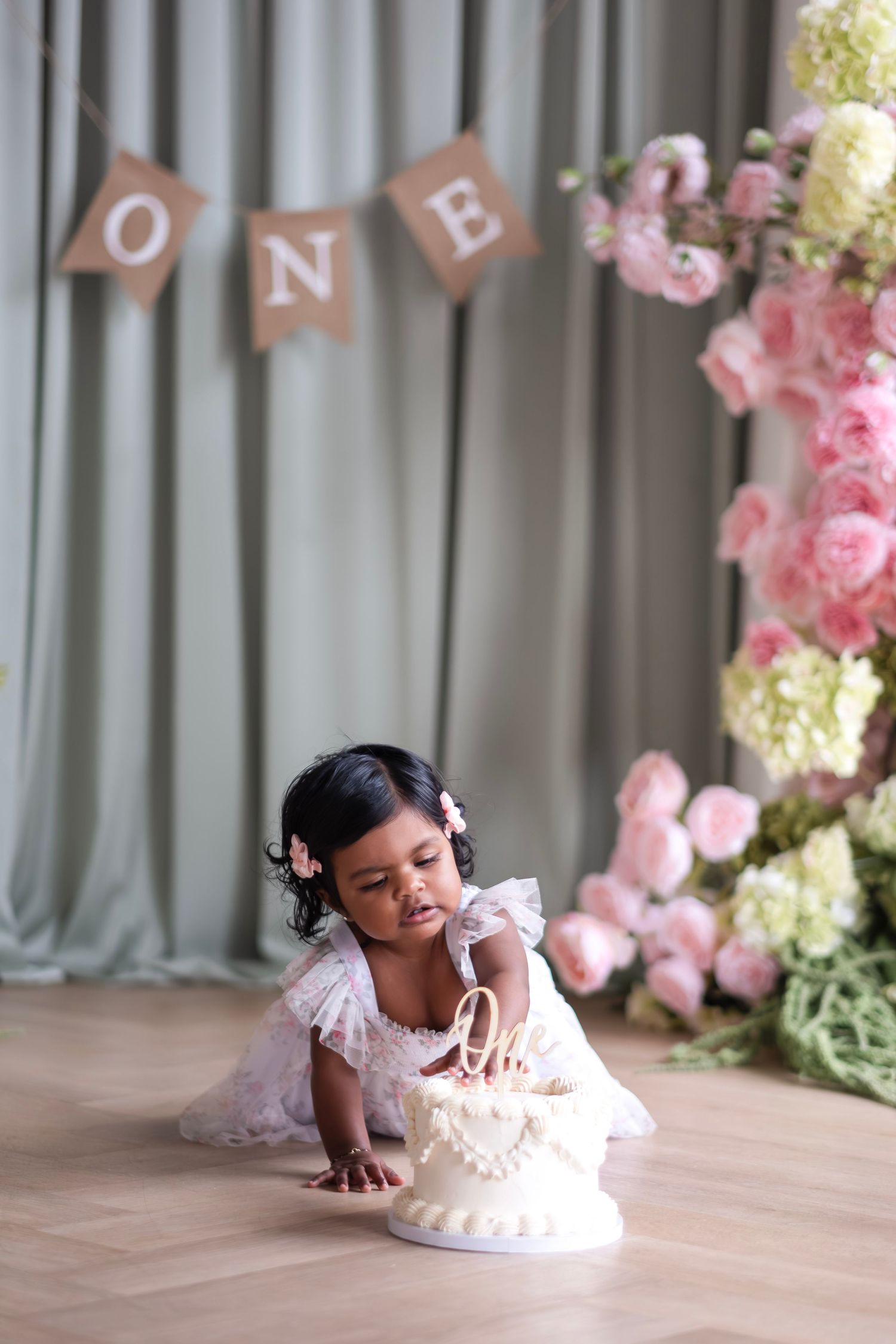A baby girl in a white dress touches her first birthday cake near a floral arrangement and ONE banner.