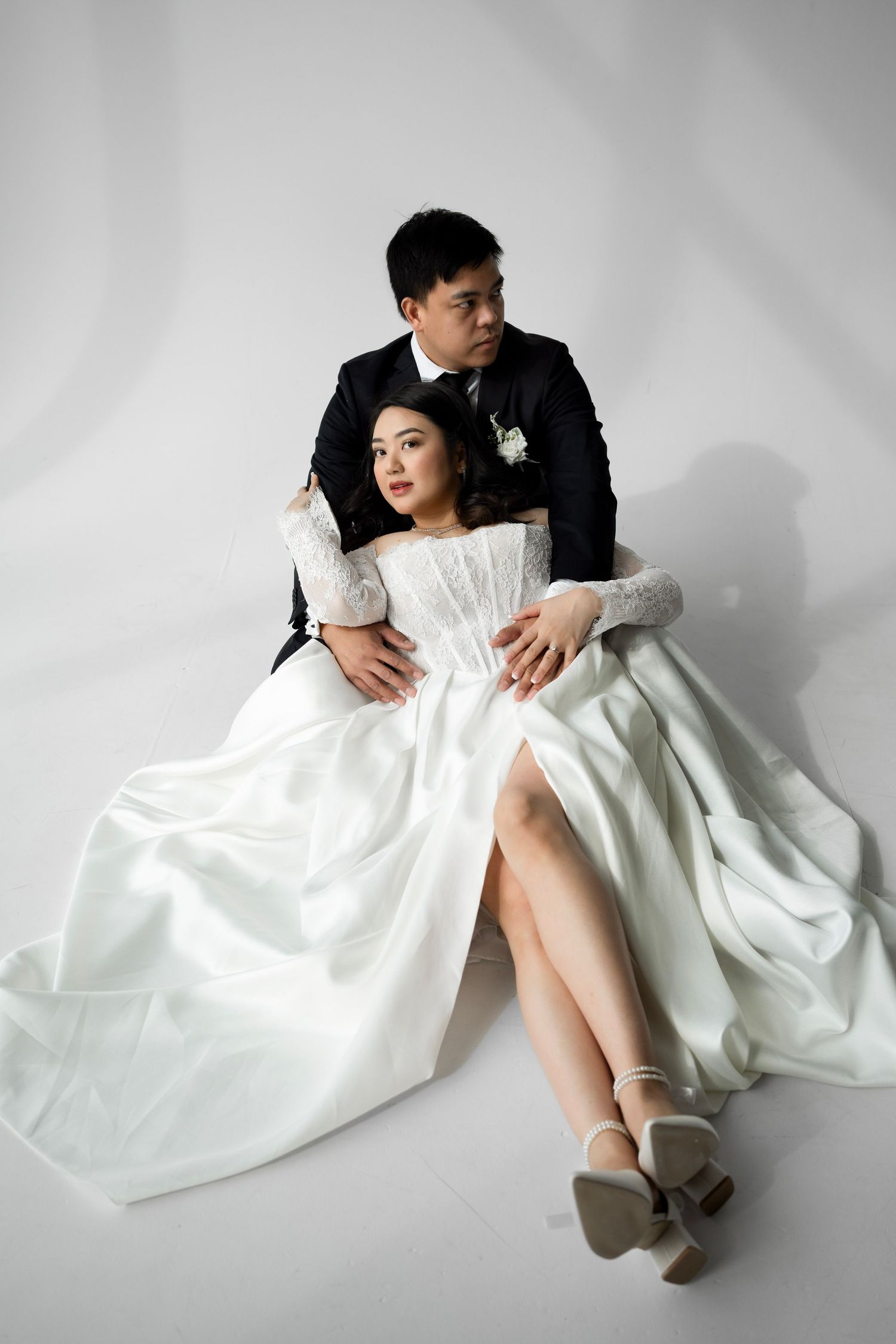 Bride and groom in wedding attire sit together on white floor, groom in black suit embracing bride in white lace gown.