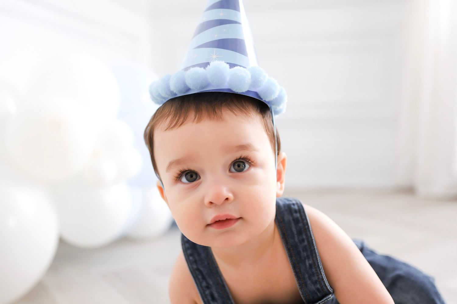 Adorable baby boy wearing a blue striped party hat and denim overalls celebrating his first birthday.