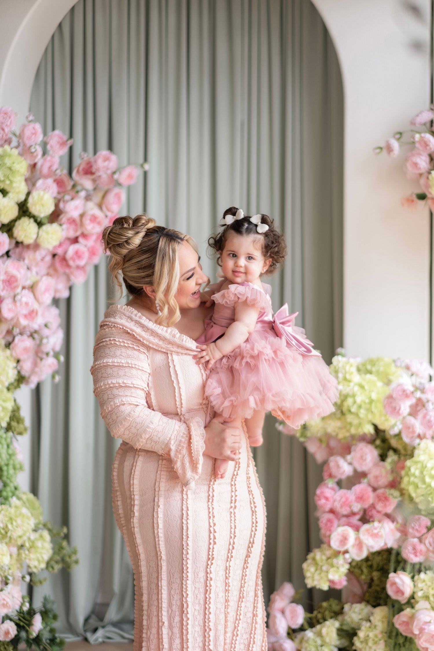 A mother in a pink gown holds her baby girl in a pink tutu dress, surrounded by lush pink and white floral arrangements.