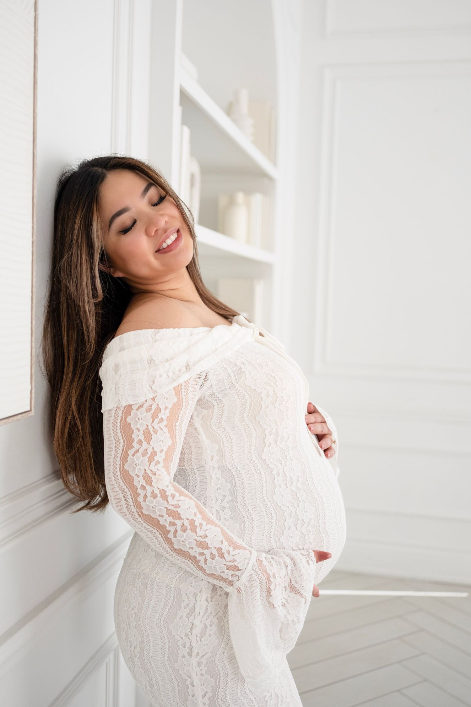 Smiling pregnant woman in white lace off-shoulder dress cradling baby bump, leaning against white wall.