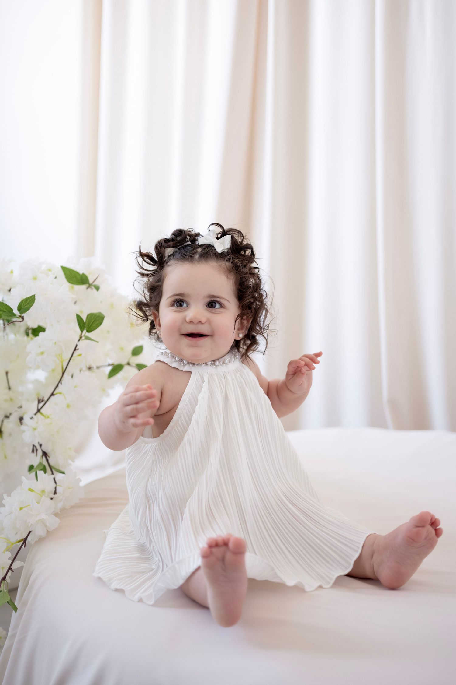 Adorable baby girl in white pleated dress with curly hair and bow sits barefoot beside white flowers.
