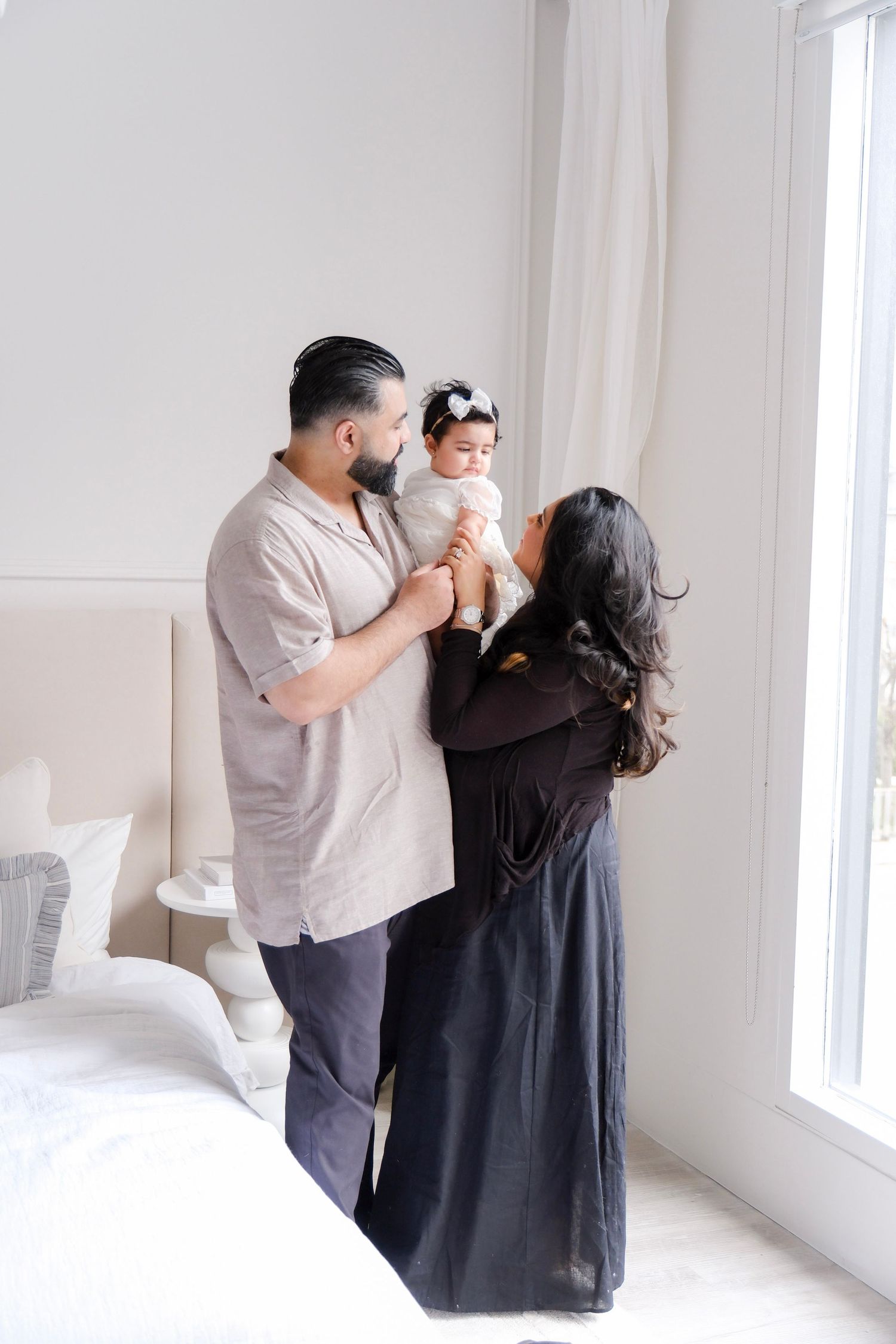 Parents in casual attire holding and interacting with small child near bright window in minimalist white room.