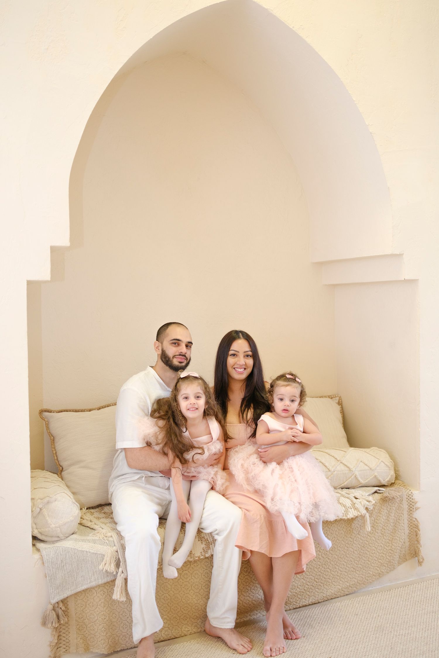 Family in coordinated white and pink outfits sitting together on beige sofa under arched alcove in bright room.