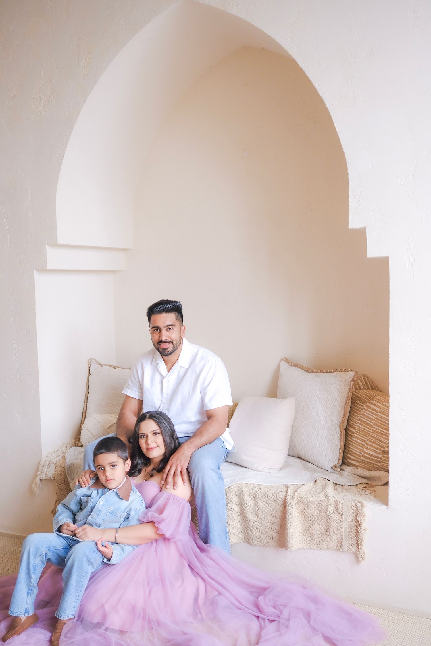 A family sits together in a white arched alcove with cushions while wearing coordinated casual outfits in blue and pink tones.