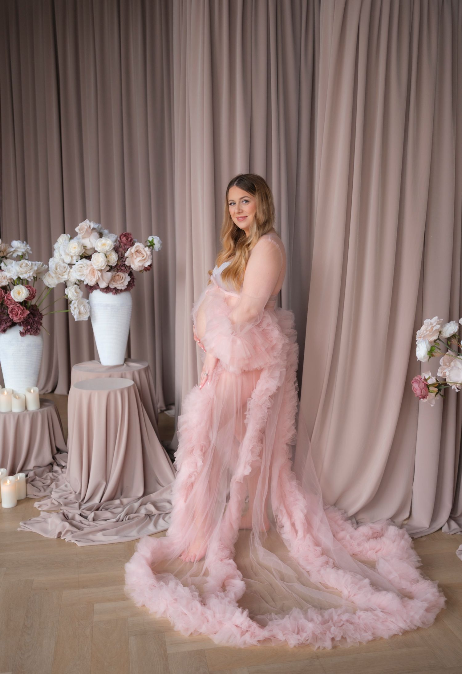 A maternity photoshoot features a flowing pink tulle gown against a rose-colored backdrop with floral arrangements.
