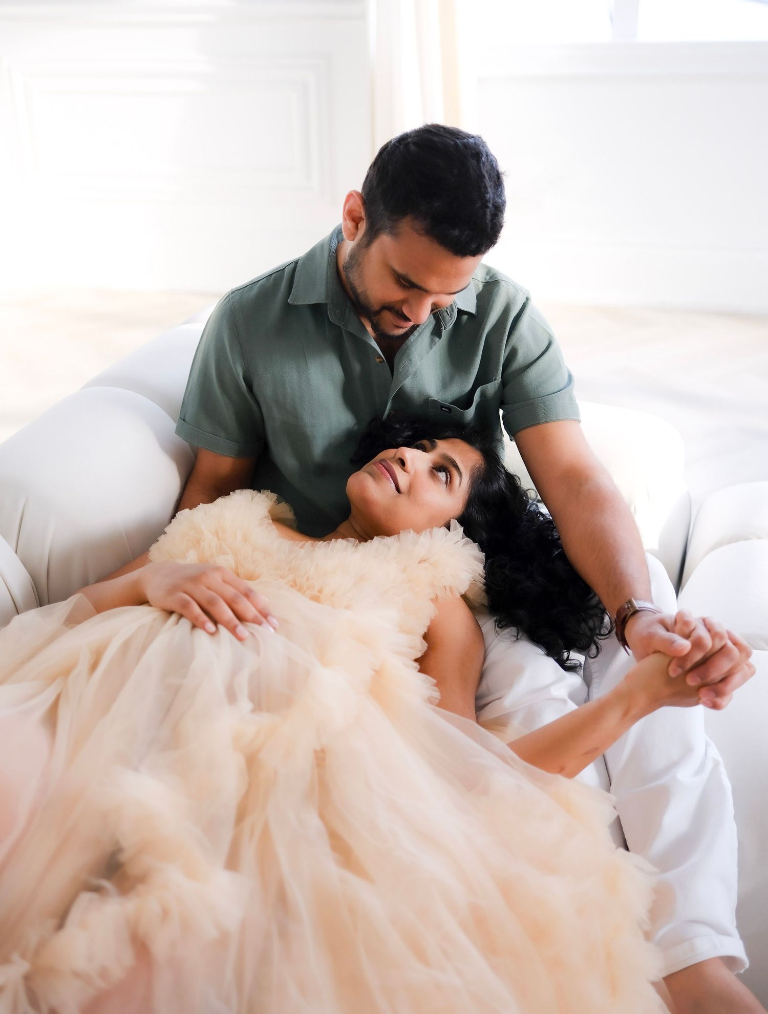 Couple shares an intimate moment together on white bedding, with her wearing a flowing blush tulle gown.