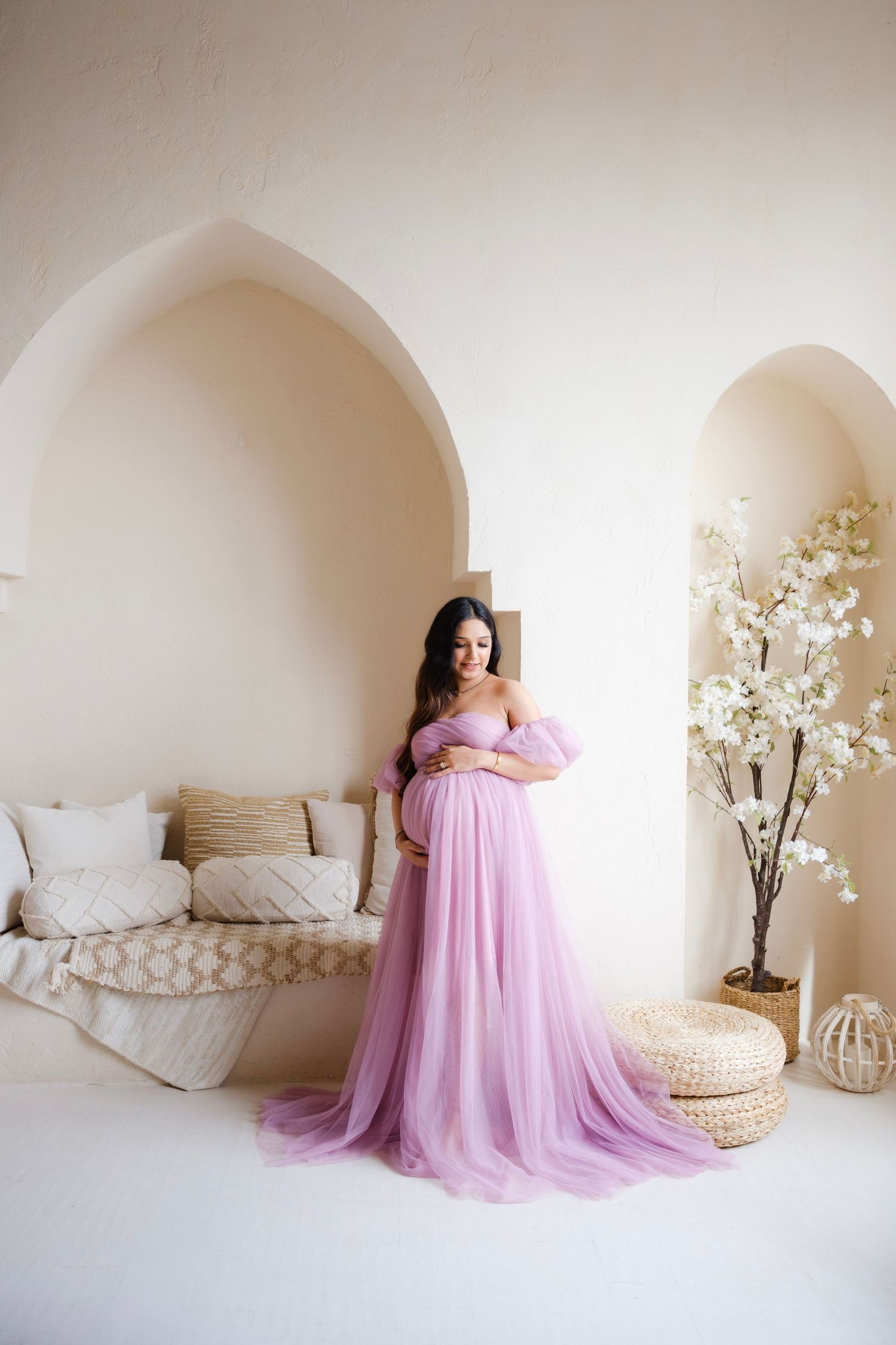 Pregnant woman in flowing lavender tulle gown poses gracefully in elegant cream interior with arched walls.