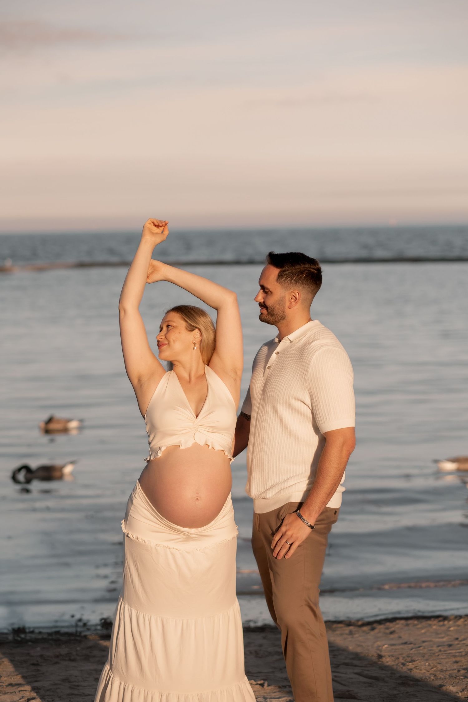 A pregnant couple poses joyfully at sunset on a beach, the woman raising her arm while showing her baby bump.