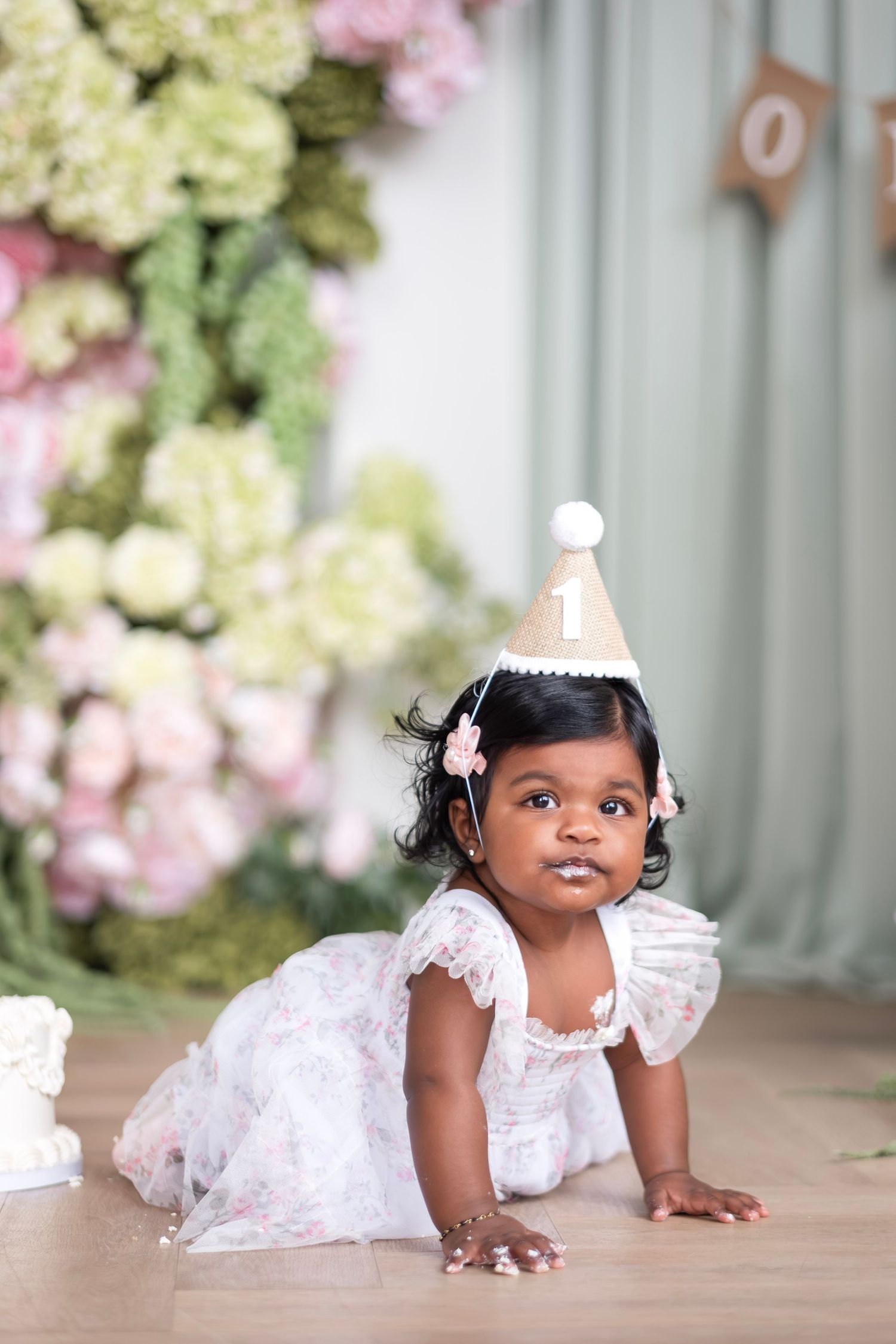 A one-year-old girl in a white dress and birthday hat crawls on the floor near a smash cake and flowers.