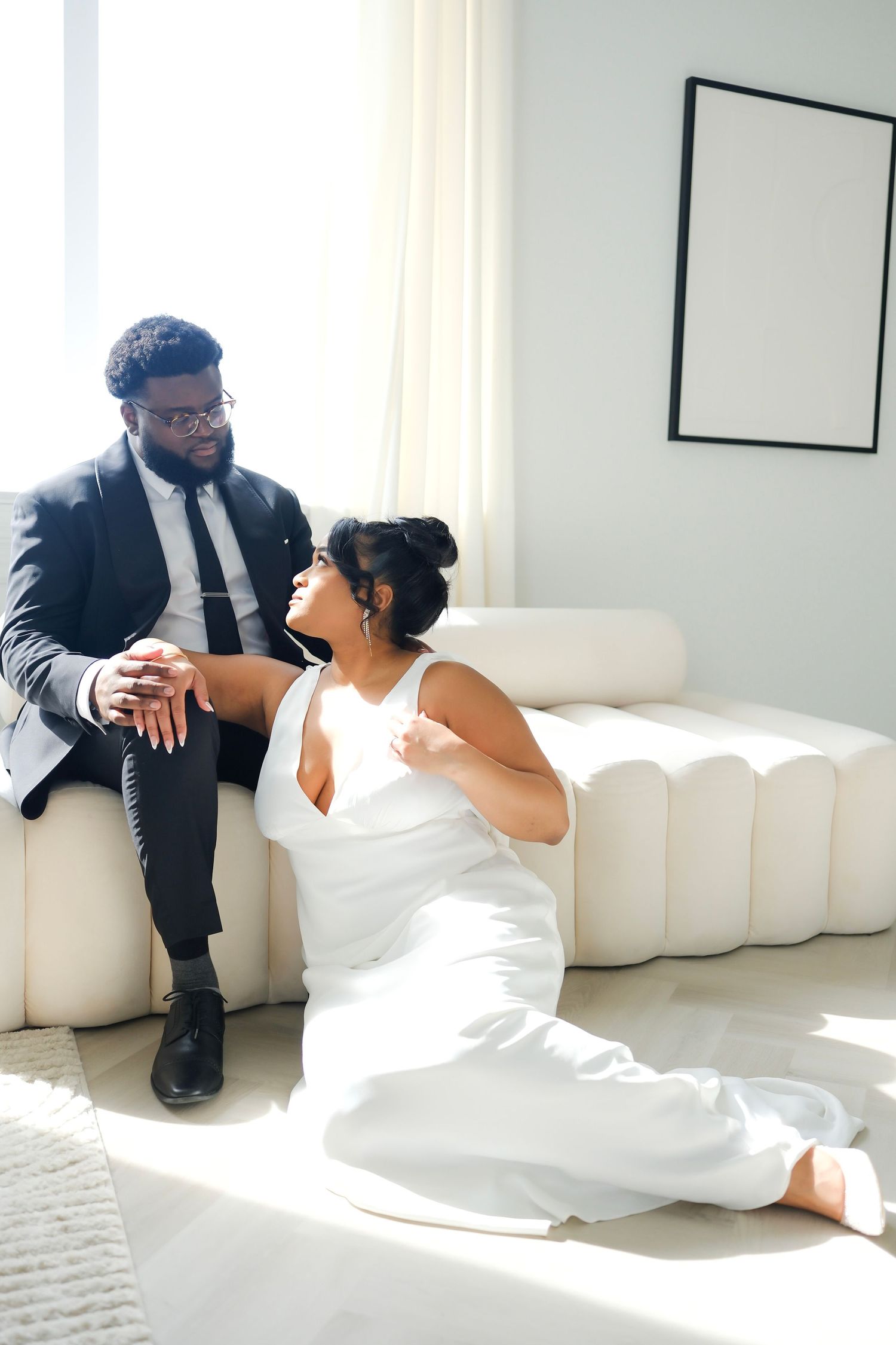 A couple in wedding attire sit together on a white modern couch in a bright minimalist room with natural lighting.