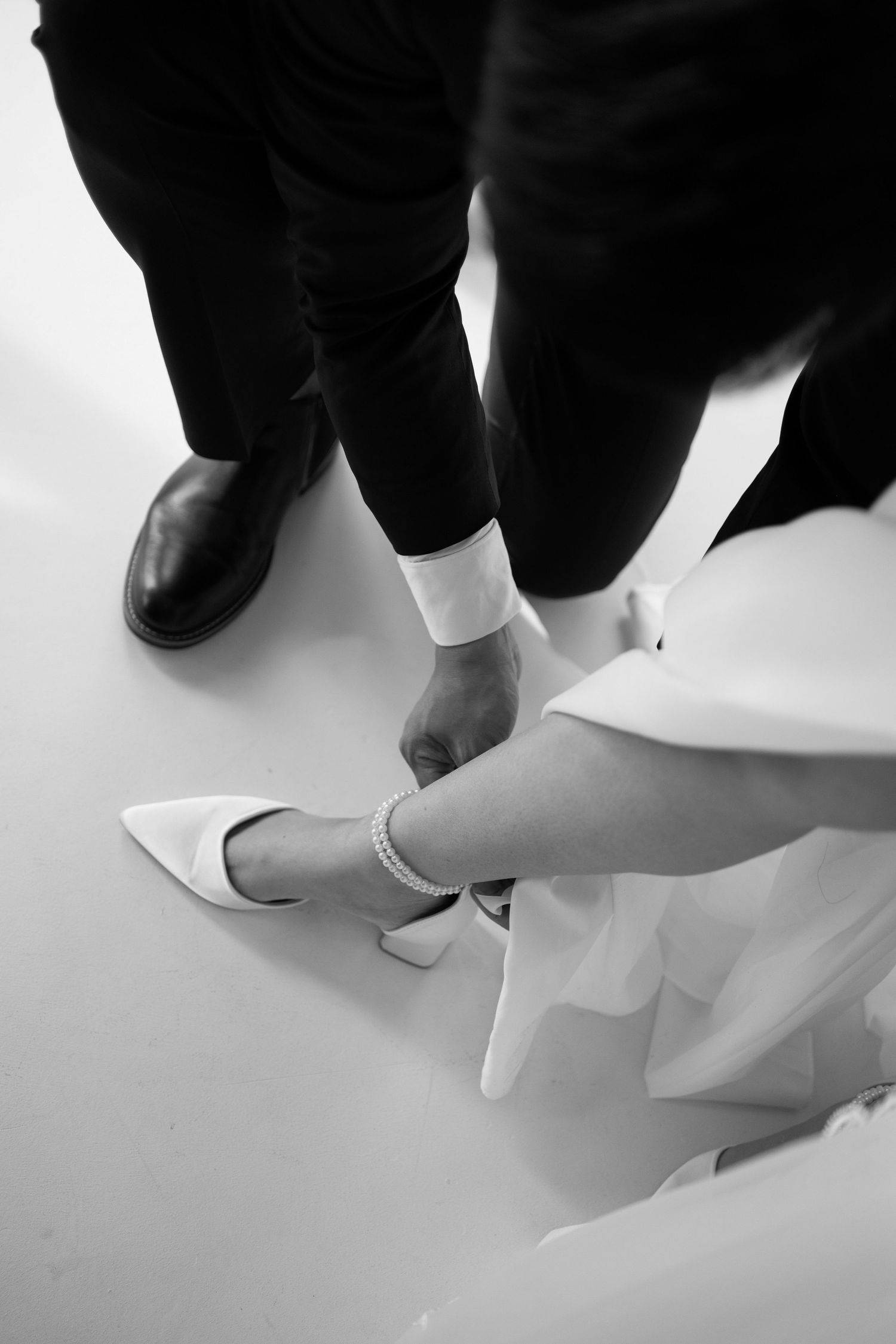 Black and white photo of groom fastening ankle strap on brides white heeled wedding shoe.