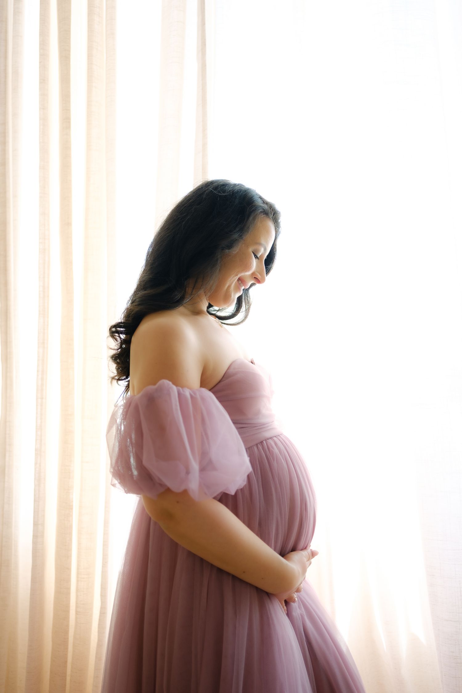 A pregnant woman in a flowing pink off-shoulder dress poses by a window in soft natural light.