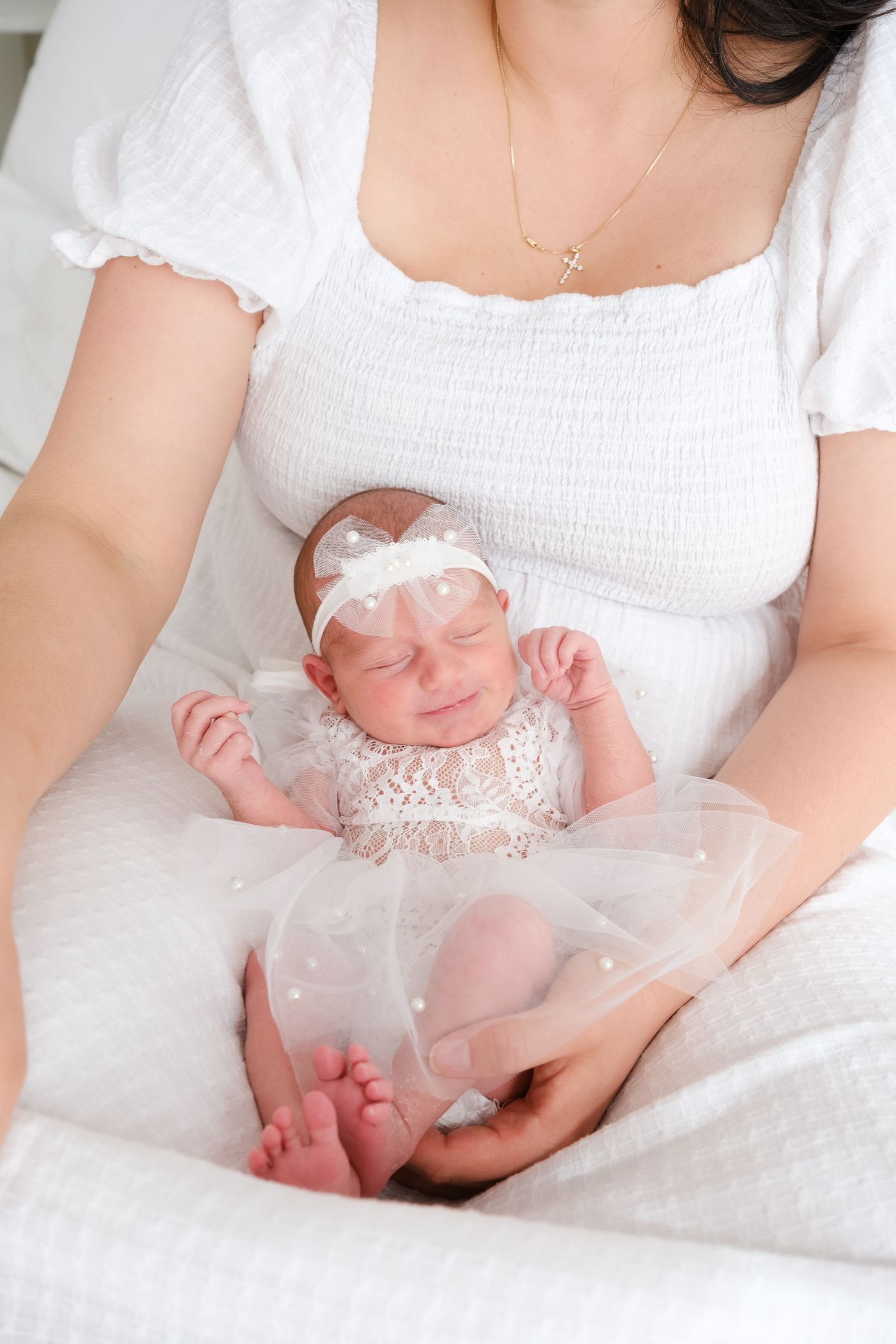 A newborn baby wearing a white lace dress and tulle bow headband rests peacefully on white bedding.