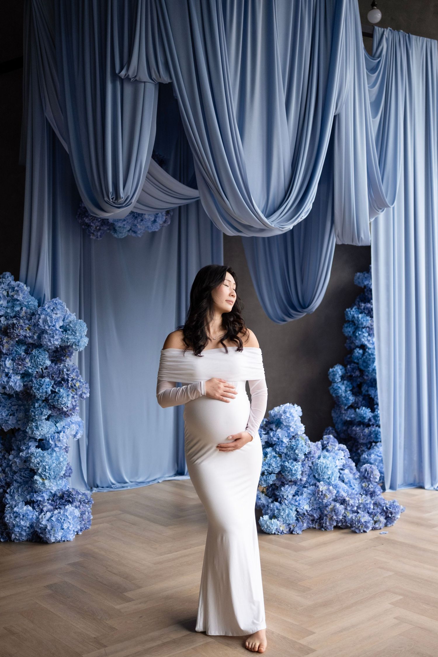 Pregnant woman in white off-shoulder gown poses with blue hydrangeas and draped fabric backdrop.