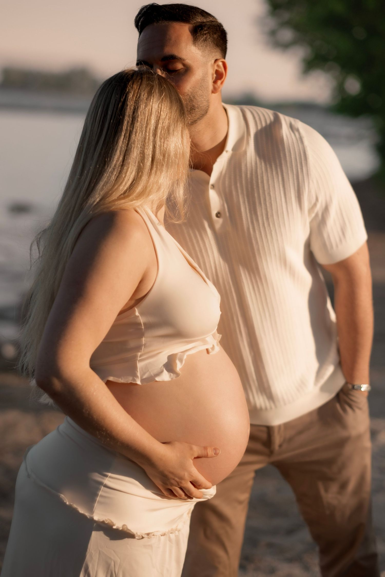 A man tenderly kisses a pregnant woman on the head while cradling her baby bump on a beach at sunset.