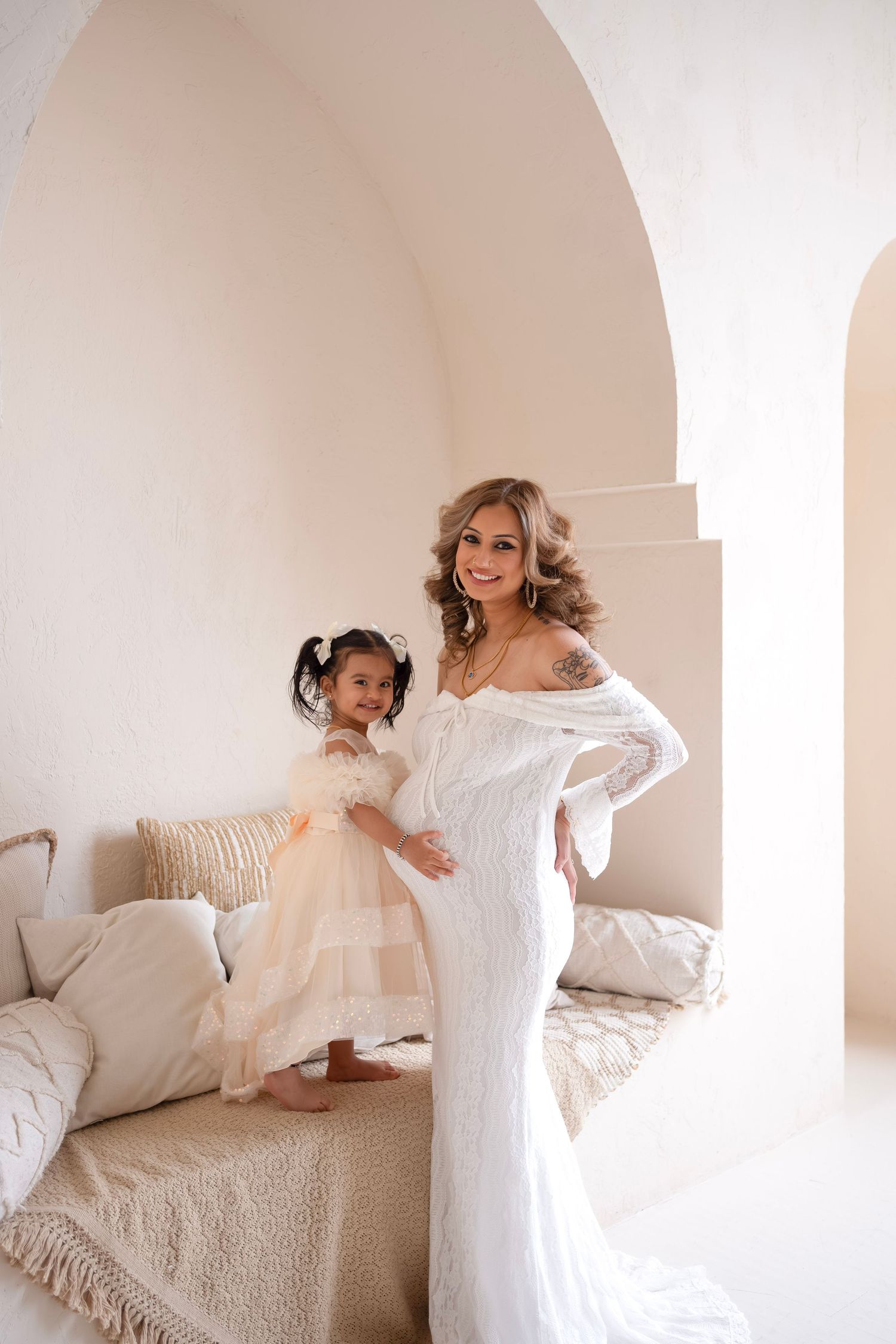 Bride in white lace gown poses with young flower girl in blush dress in elegant arched white interior.