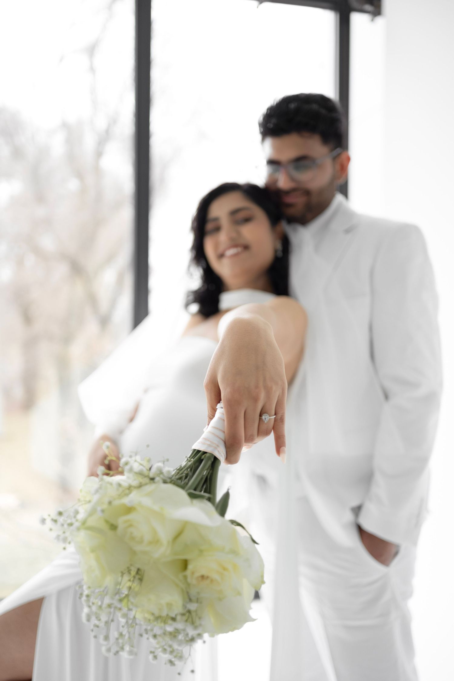 Smiling bride in white dress showing diamond ring and bouquet of white roses while groom embraces her from behind.
