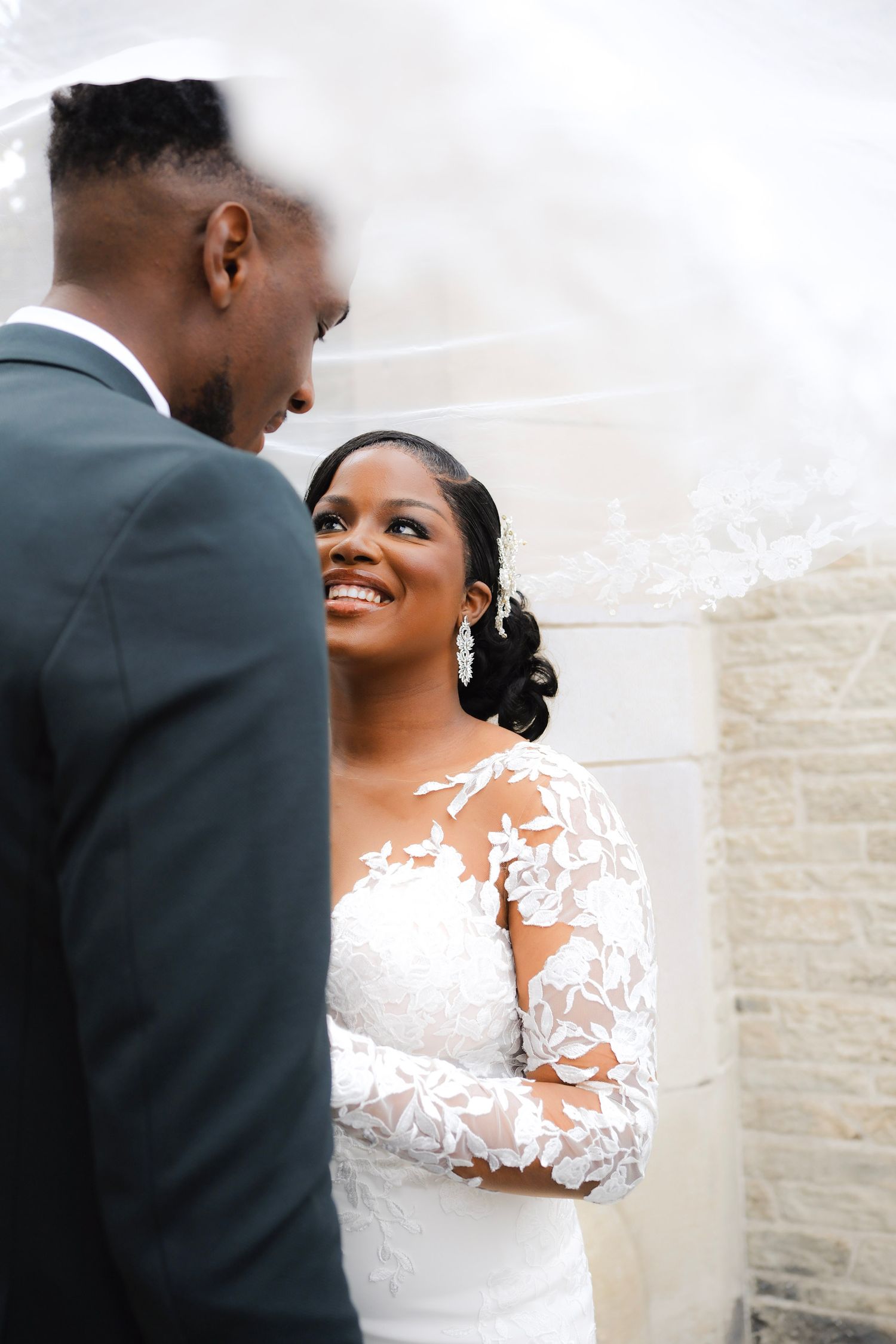 A joyful newlywed couple share a romantic moment in their wedding attire against a light-colored wall.