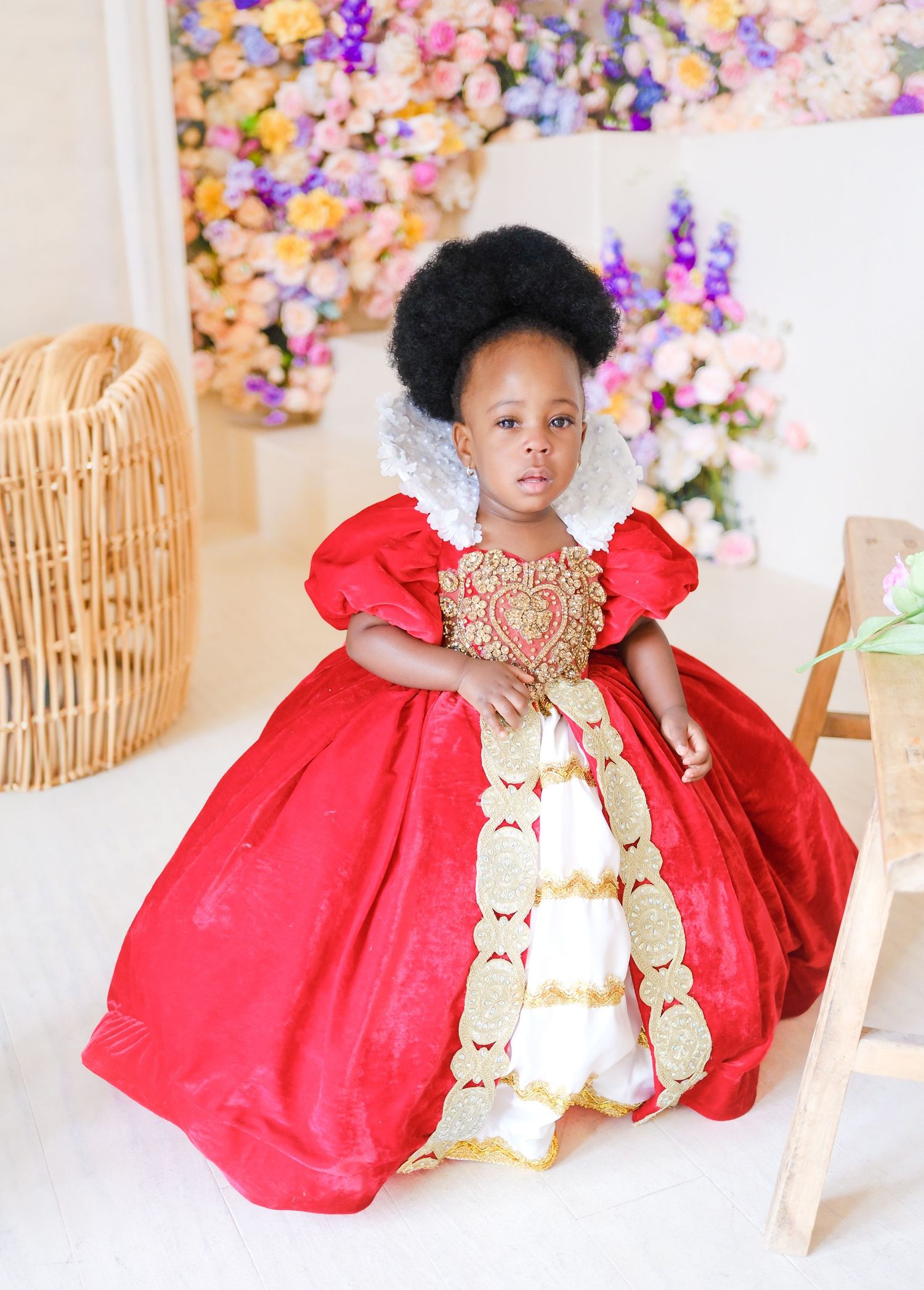 A small child wearing an elegant red and gold princess ball gown poses in front of a floral backdrop with a woven basket nearby.