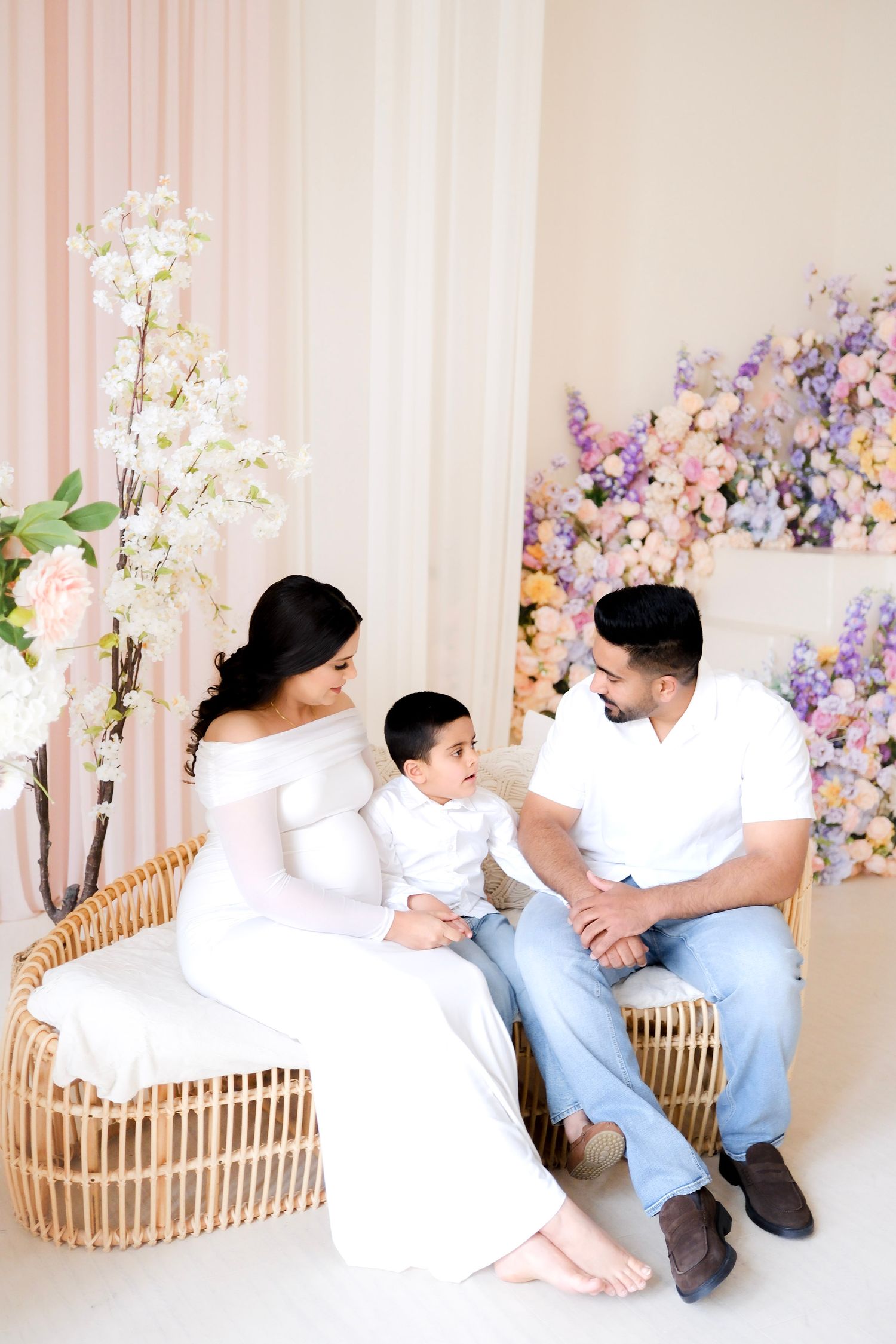 Family sitting together on rattan bench with elegant white and purple floral decorations in light studio setting.