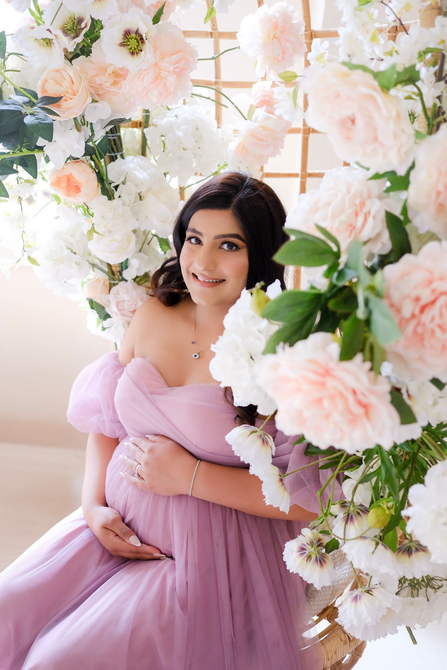 A romantic maternity portrait features a lavender off-shoulder gown against a backdrop of peach and white floral arrangements.