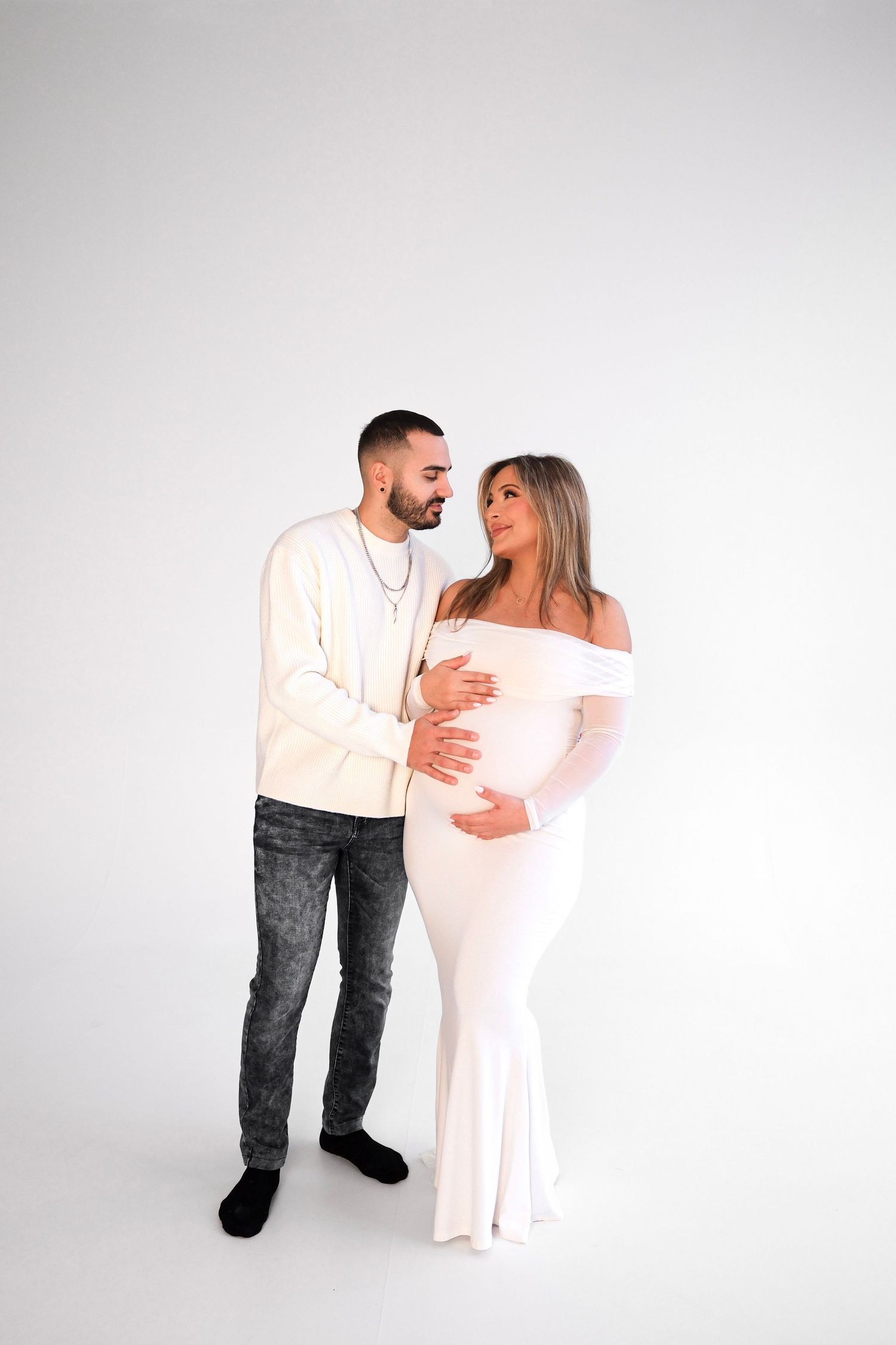 A couple shares an intimate moment during a maternity photoshoot against a white background.