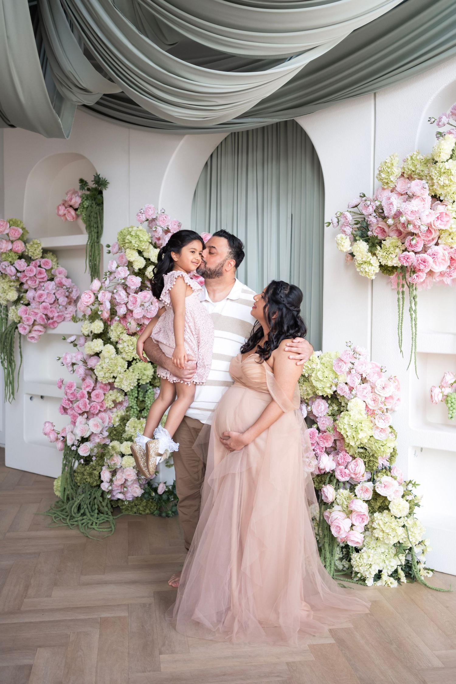 A family maternity photo with a pregnant woman in a pink gown, man holding a girl, surrounded by lush floral arrangements.