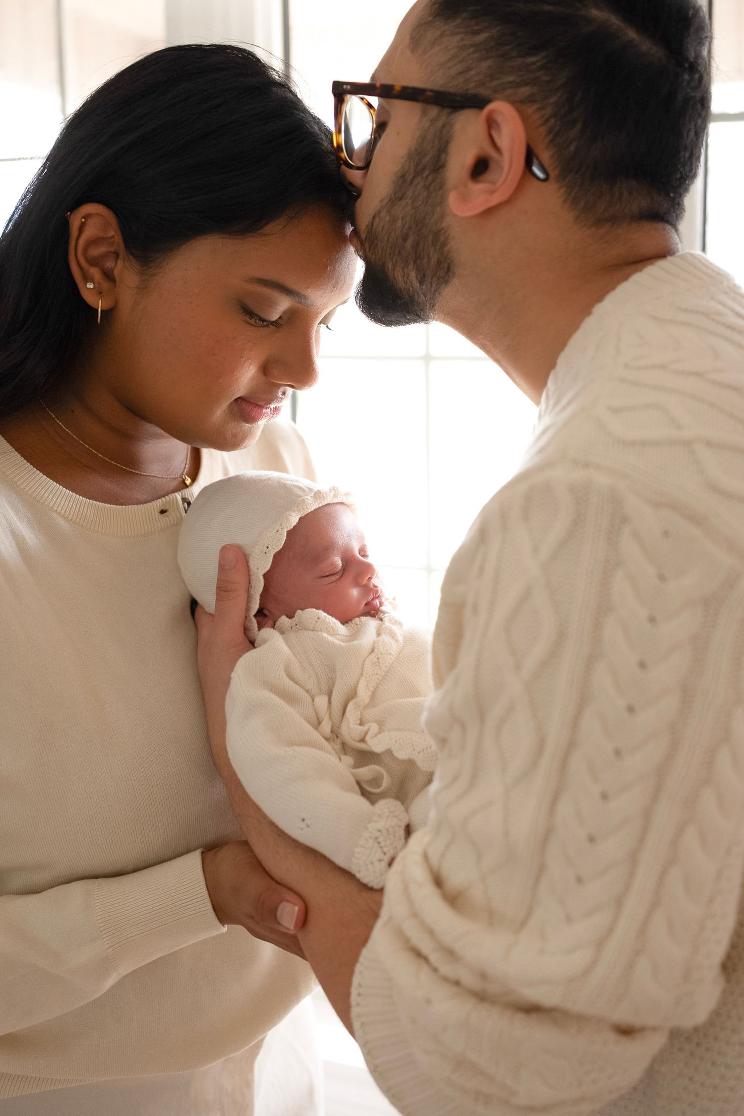 New parents in white sweaters lovingly cradle their sleeping newborn baby wrapped in white.