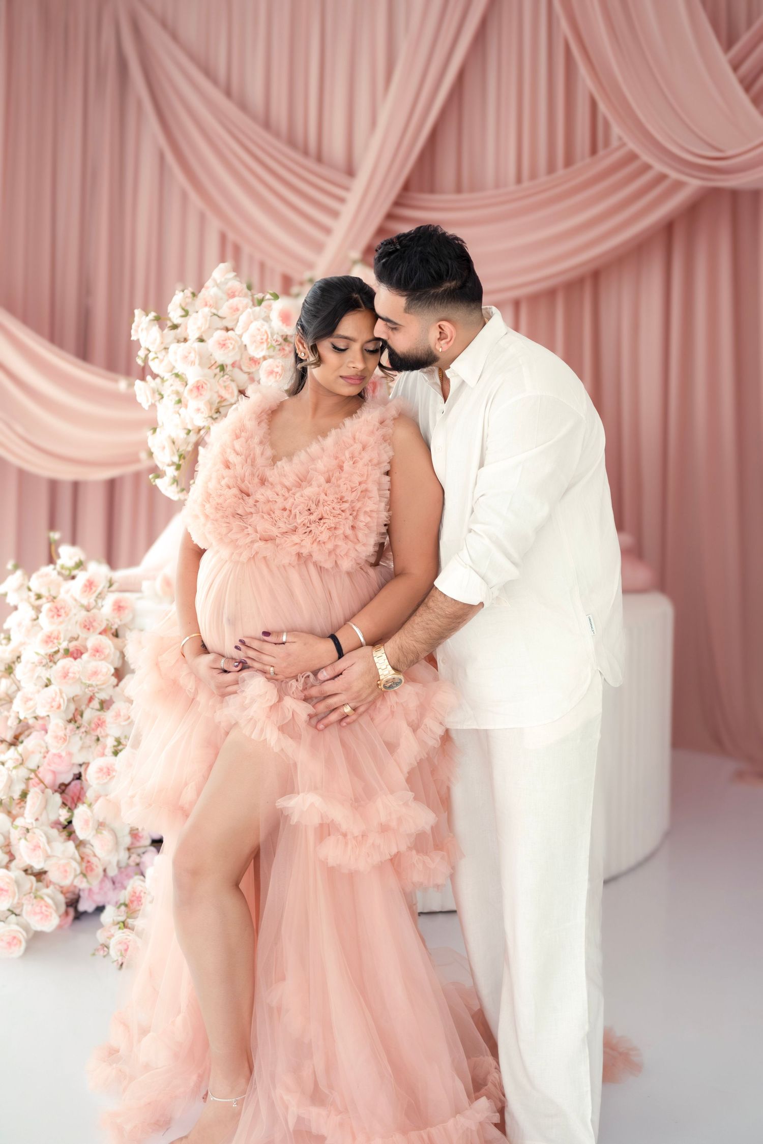 A romantic maternity photoshoot with pink draped backdrop featuring a couple in white and blush pink outfits.