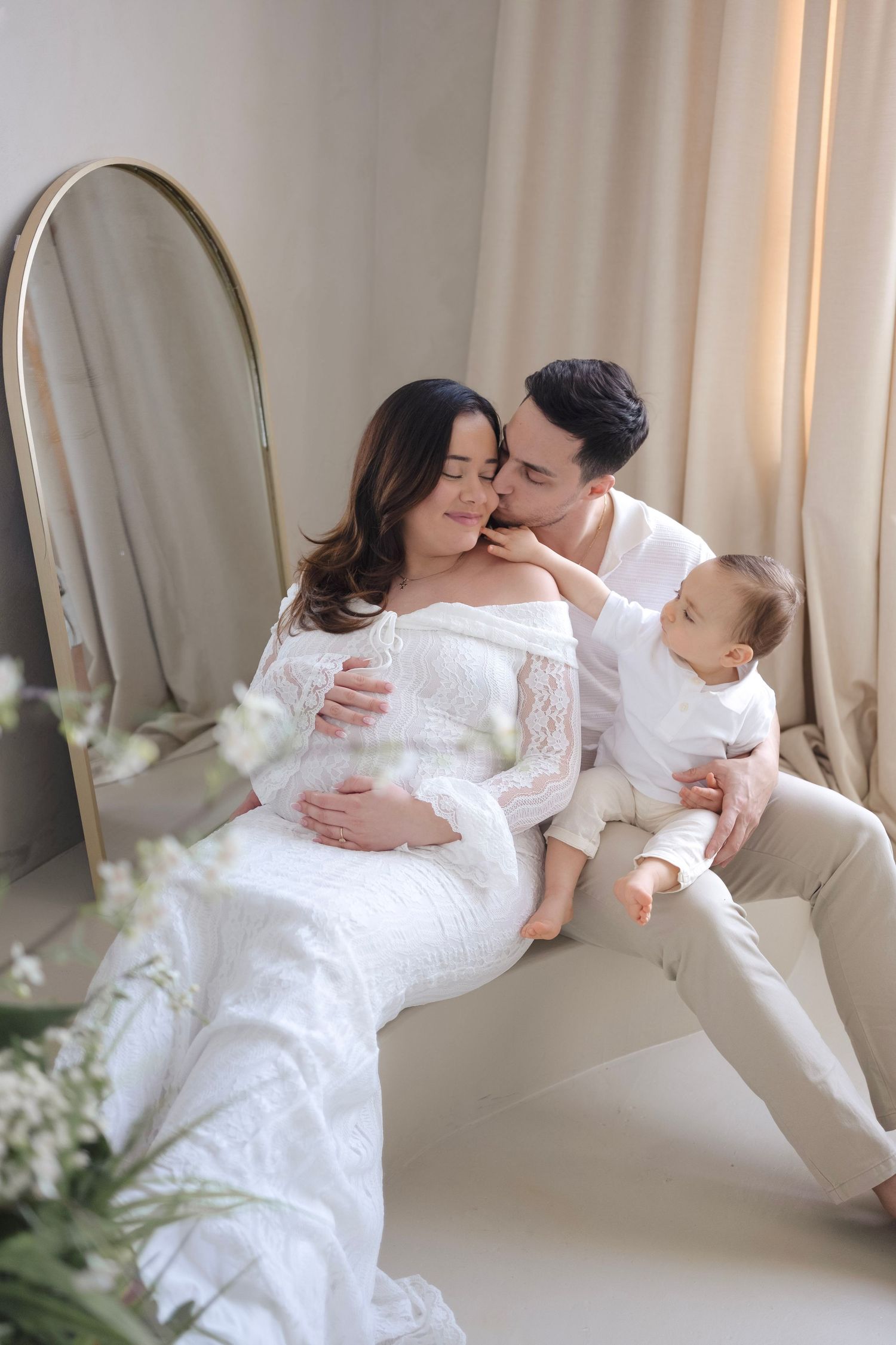 Pregnant woman in white lace gown with husband and baby in bright studio maternity photo session.