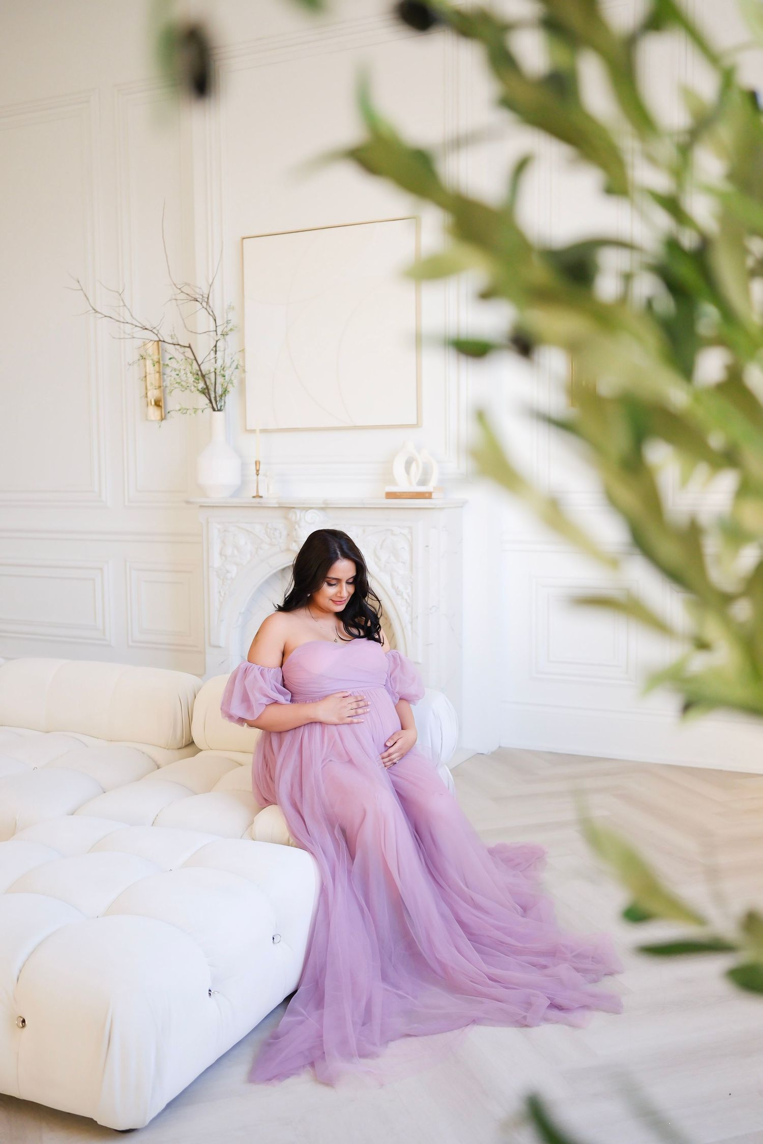 Pregnant woman in flowing purple dress sits elegantly on white couch during maternity session. Toronto, maternity, photographer,