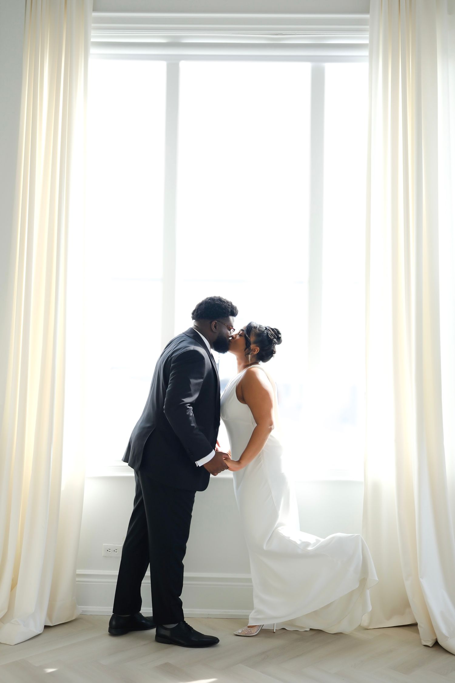 Newlywed couple in black suit and white dress share an intimate silhouette moment by a bright window with cream curtains.